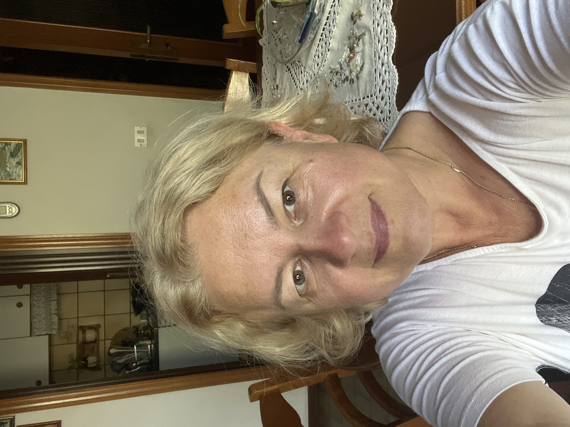 In the city of Ostra Vetere, Italy, an image captures a woman in her 50s with blonde, shoulder-length hair and hazel eyes. She is wearing a white t-shirt and a gold necklace. The woman is taking a selfie in what appears to be a home setting. In the background, a doorway leads to a kitchen, visible through an open door. The kitchen features beige tiled walls and white cabinets. To the right of the woman, a dining table is set with a white, crocheted tablecloth and a glass bowl. Wooden chairs are also visible. The lighting suggests it is daytime.