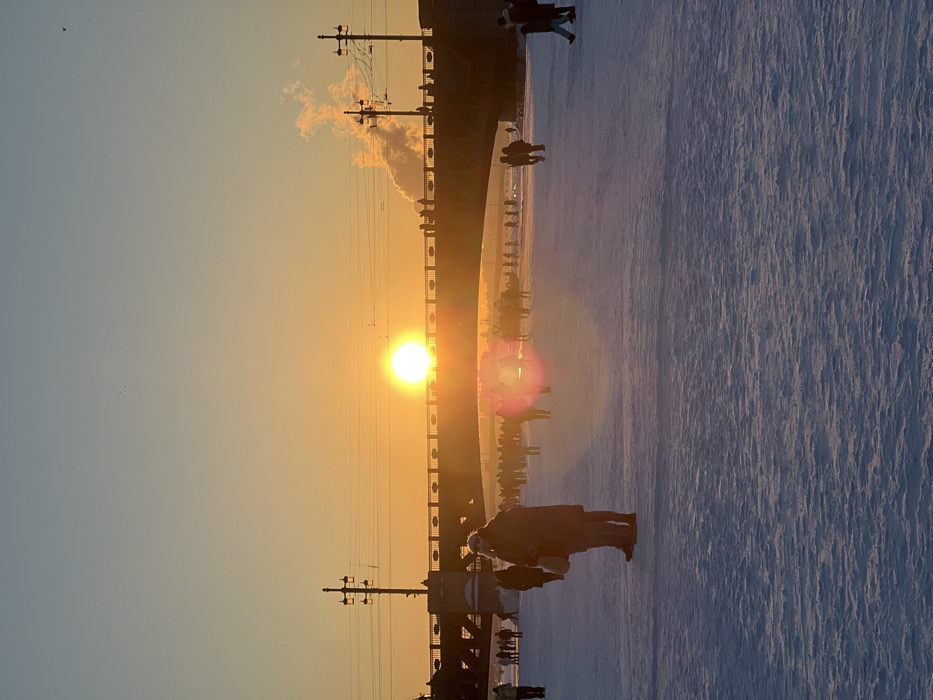 The image depicts a winter scene on the frozen Neva River in Saint Petersburg, Russia, with the Troitsky Bridge spanning horizontally across the middle ground. The sun, a bright orange-yellow orb, is positioned low on the horizon, slightly left of center, directly above the bridge, indicating late afternoon. A prominent magenta-pink lens flare emanates from the sun in the lower-middle frame.
The environment consists of a vast expanse of uneven, textured snow and ice covering the frozen river in the foreground and midground. The sky above transitions from a pale blue to a warm orange-yellow near the horizon.
Numerous individuals are present. In the left-middle foreground, two heavily clothed figures stand with their backs to the viewer, observing the scene. A large crowd of people, appearing as dark silhouettes against the bright snow and sun, are scattered across the frozen river in the midground, walking or standing.
The Troitsky Bridge, a dark-colored metal truss structure, features several tall, ornate lampposts with overhead electrical lines. Silhouetted figures, representing pedestrians and potentially vehicles, are visible moving across the bridge deck. To the right of the bridge, a distinct plume of white smoke or steam rises into the orange sky from an unseen source beyond the bridge.