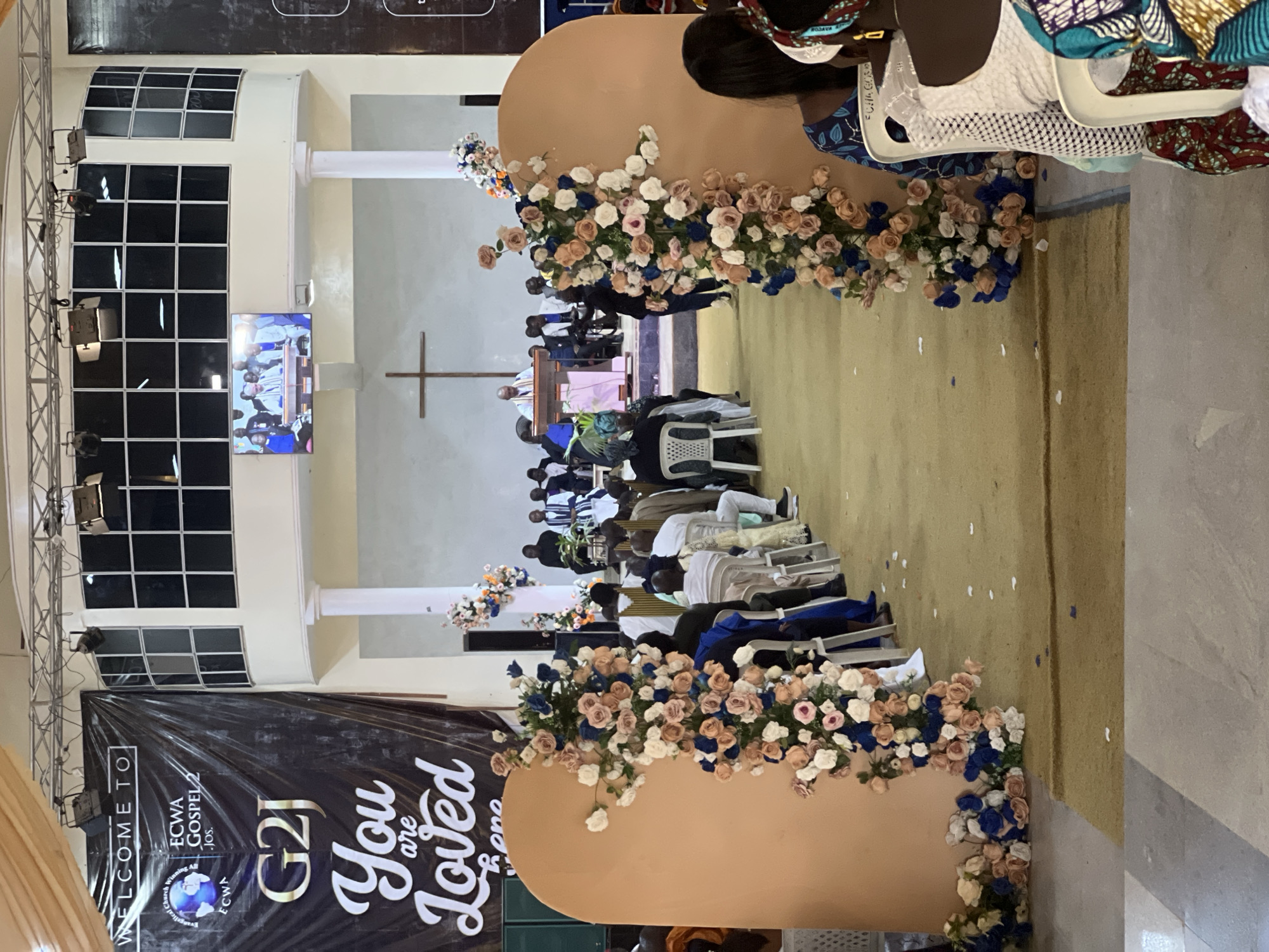 The image captures an indoor event, likely a church service or ceremony, within the ECWA Gospel Church in Kabong, Nigeria. The focal point is a decorated aisle with floral arrangements featuring peach, white, and blue roses on large, peach-colored archways. A banner at the back of the venue reads "WELCOME TO ECWA GOSPEL G2J You are Loved here."
The scene is populated by attendees seated in chairs, facing a raised platform where a speaker is addressing the congregation. A large screen displays a live feed of the speaker. The environment appears to be a church hall with a carpeted floor and a stage area. The lighting suggests an artificial light setup, common for indoor events. The audience members are diverse in age and attire, with some individuals dressed in formal wear and others in traditional Nigerian clothing. The overall atmosphere seems to be one of solemnity and communal gathering.