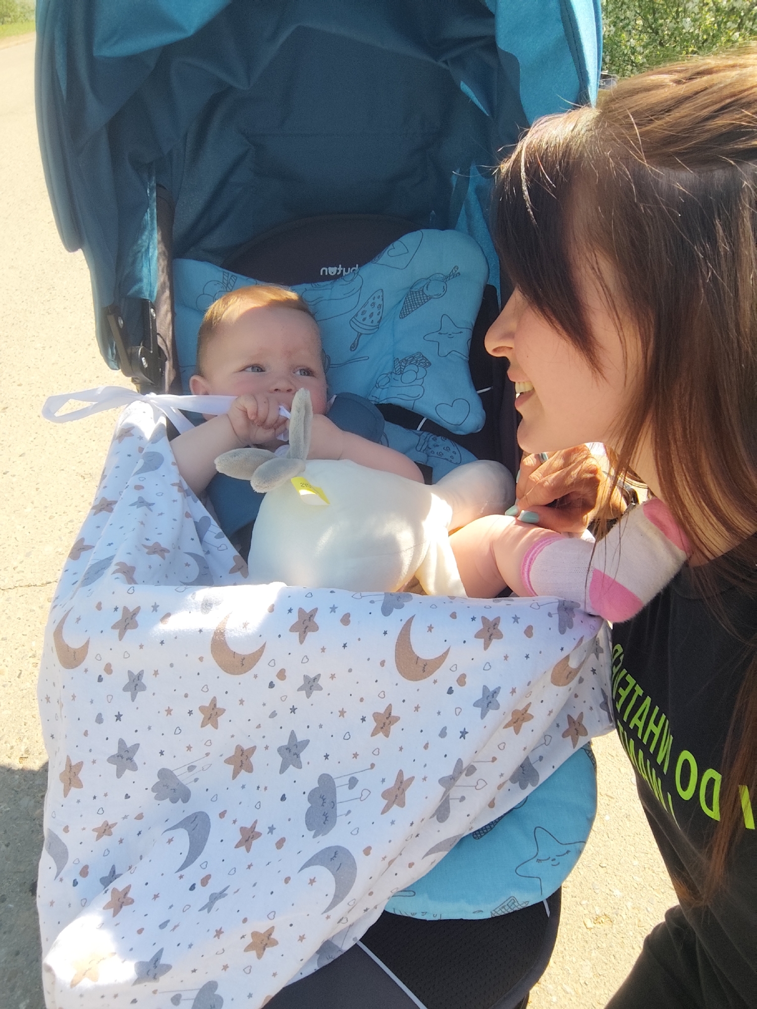 A baby is seated in a blue stroller with a white blanket adorned with celestial designs. The baby has a pacifier in its mouth and a white stuffed animal with gray ears nearby. A woman with brown hair stands beside the stroller, looking at the baby with a smile. The stroller is on a paved surface outdoors. The letters "DO WHAT" are visible on the woman's black shirt.