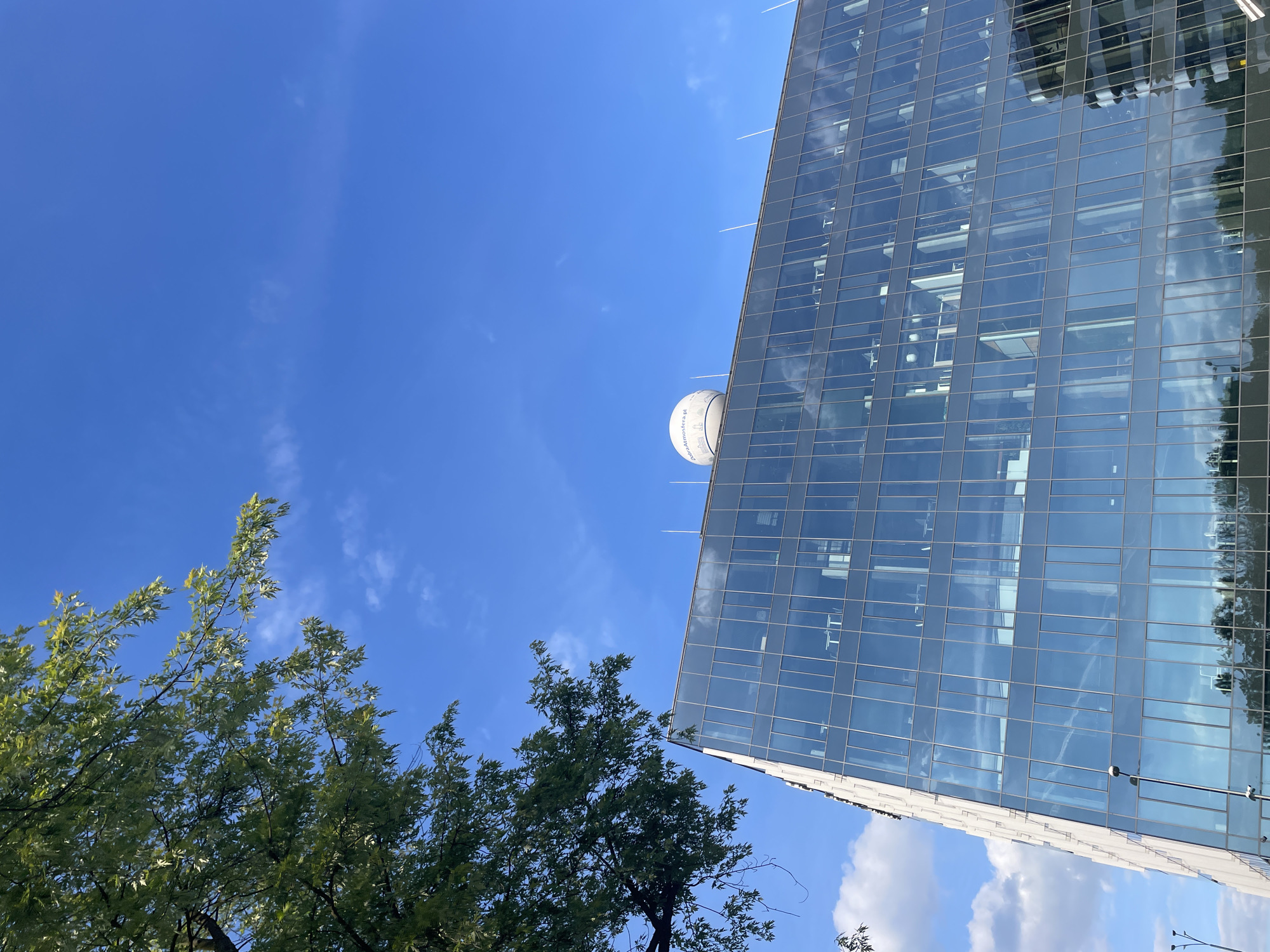 The image captures a low-angle view in an urban setting. A tall, modern building with a glass facade dominates the right side of the frame. The glass reflects the sky and nearby trees. A large, white, spherical object is positioned near the top of the building. Trees with green foliage occupy the lower-left portion of the image. The sky is blue with wispy clouds. The location is Unknown, Unknown.