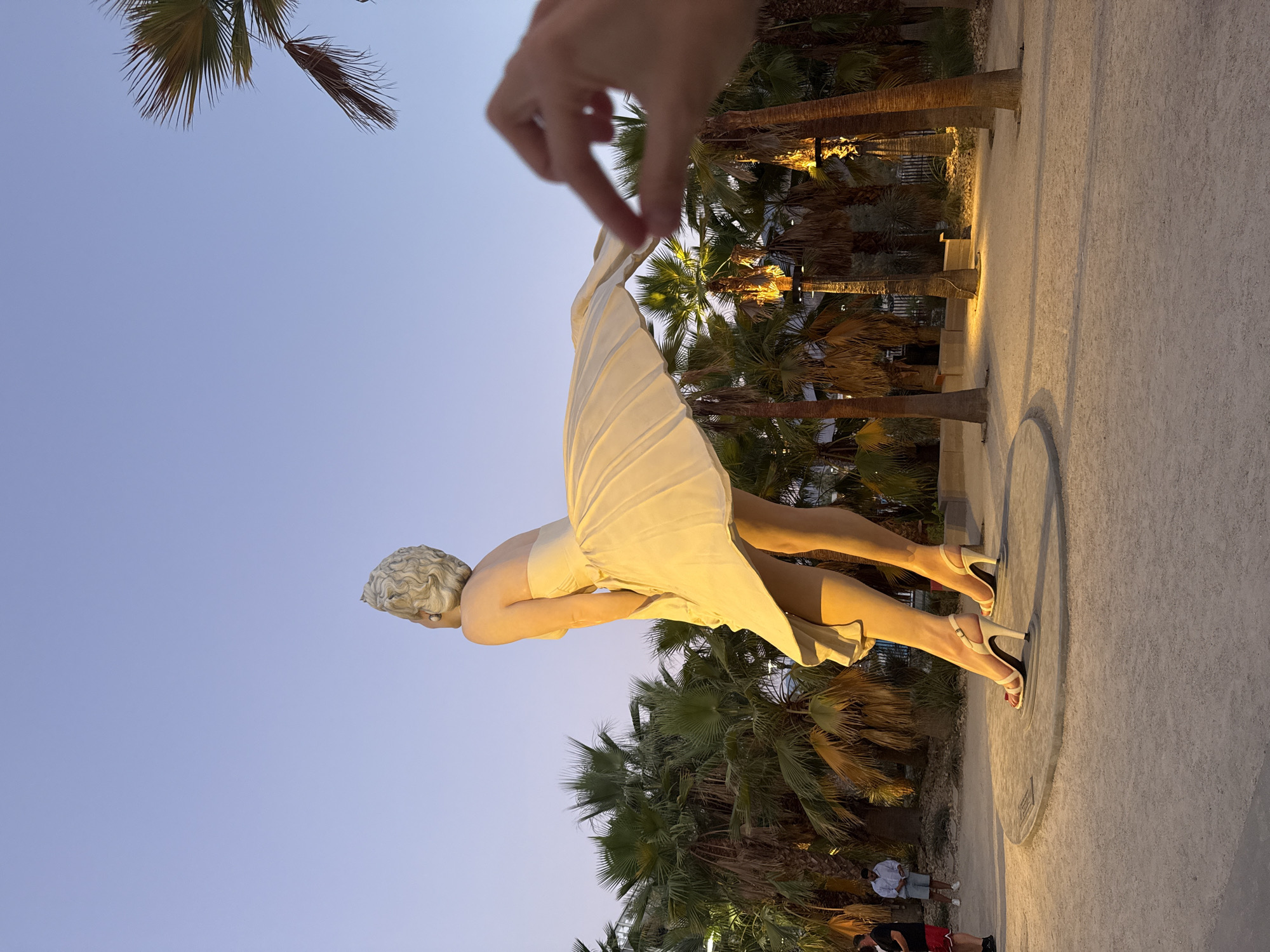The image captures a nighttime outdoor scene featuring a large statue of Marilyn Monroe in her iconic "The Seven Year Itch" pose. The statue is illuminated from below, highlighting her billowing white dress and legs. The location is the Marilyn Monroe Statue in Palm Springs, California, United States of America, a popular tourist attraction. Palm trees surround the statue, with some of them also lit. In the foreground, a hand is positioned to appear as if it's holding the hem of the statue's dress. Two men are visible in the background, one standing near the statue and another further to the left, observing the scene. The ground is a light-colored gravel or sand. The sky above is a clear, deep twilight blue.