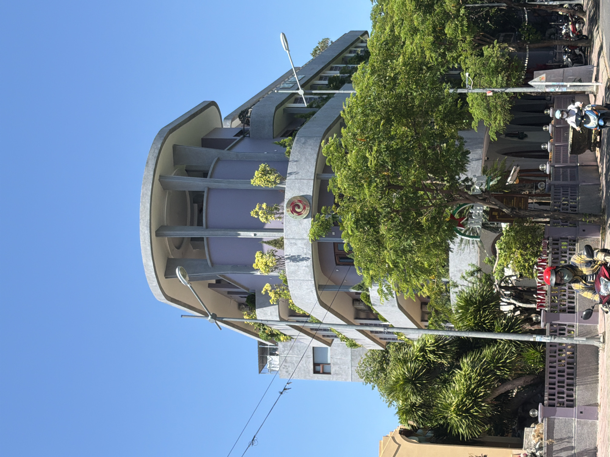 In Nam Nha Trang Ward, Vietnam, a modern, multi-story building with a distinctive curved facade dominates the scene. The building features multiple balconies with lush green plants and trees, creating a vibrant contrast with its gray and lavender exterior. A prominent circular logo with a red and yellow swirl is visible on the building's facade. A street with a clear blue sky above is in the foreground, with a street light and power lines running across the frame. Two individuals on motorcycles are present on the street; one in the foreground wears a yellow and black plaid shirt, a red helmet, and a face mask, while another further down the street wears a white shirt and helmet. Several other motorcycles are parked along the street. The area is lined with a decorative purple fence and more greenery, suggesting a commercial or residential streetscape.