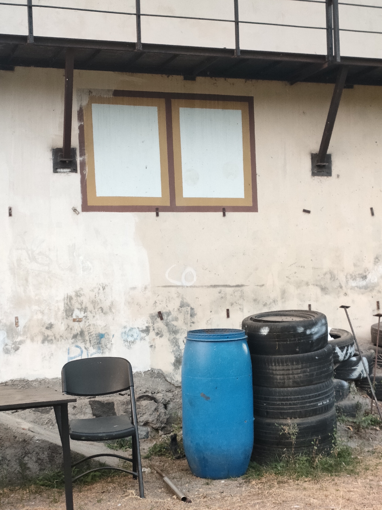 The image depicts an outdoor scene in Banguntapan, Indonesia, featuring a weathered building wall with two framed window-like areas. In the foreground, a stack of used tires is visible, alongside a blue plastic barrel. To the left, a black folding chair sits next to a table. The ground is covered with gravel and dry vegetation. The wall shows signs of age and graffiti. A metal railing runs along the top of the building. No people are present in the frame.