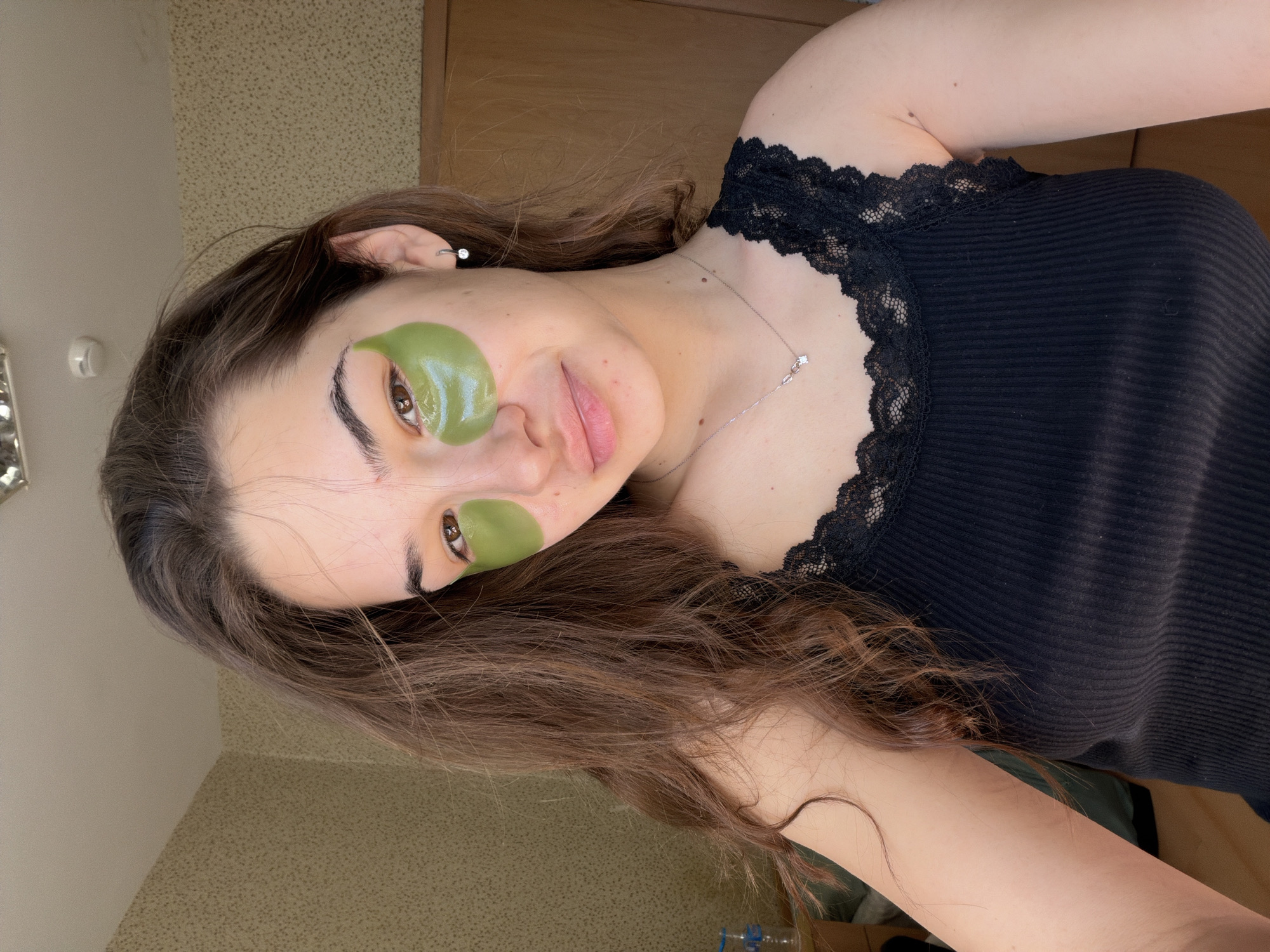 The image displays a close-up, rotated view of a young woman engaged in a self-care routine. She has long, wavy dark brown hair and fair skin. On her face, she wears two distinct green, gel-like under-eye patches; one is placed directly below her left eye, and the other slightly lower and to the right, near her nose and cheekbone. She looks calmly towards the camera with a subtle, relaxed smile.
The woman is wearing a black, ribbed tank top featuring a delicate black lace trim along the neckline. A thin silver necklace with a small, clear pendant hangs around her neck, and a small silver earring with a clear stone is visible in her left ear. Her eyes are light brown, and her eyebrows are dark and well-defined.
The background indicates an indoor, domestic setting. Behind her head is a light-colored wall with a textured pattern, next to a brown wooden surface. A small, white object, possibly a light switch or thermostat, is visible on the wall in the upper left corner. A clear plastic bottle is partially visible at the bottom right of the frame. The overall impression is one of quiet relaxation and personal well-being. No discernible text is visible in the image.