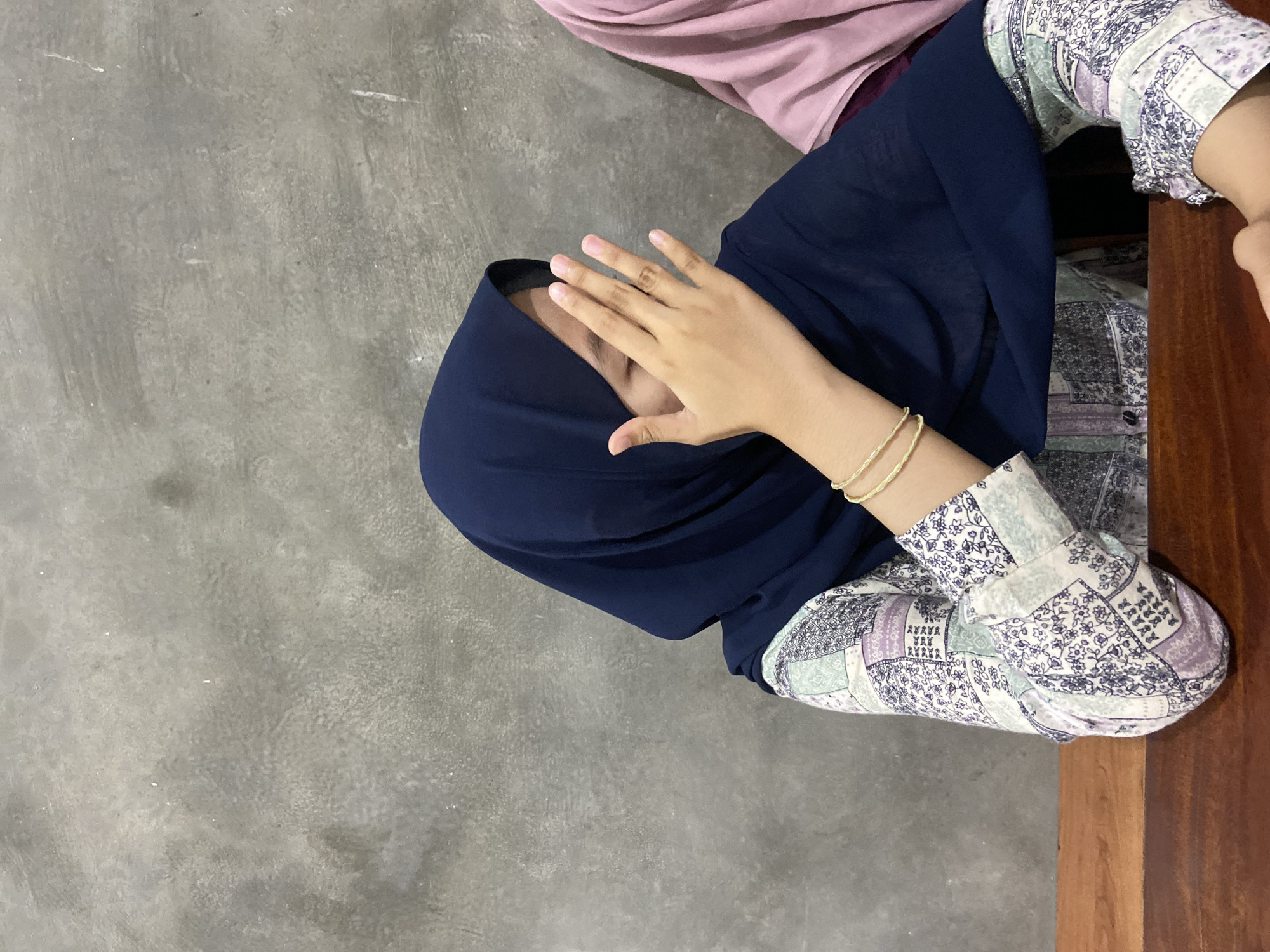 In Sekar Mawar, Indonesia, an image captures a person wearing a navy blue hijab and a patterned long-sleeved shirt. The person is partially covering their face with their right hand, showing their fingers and fingernails. They are wearing two thin gold bracelets on their left wrist. The background is a textured, gray wall. A wooden surface, likely a table or bench, is visible in the foreground. Another person wearing a pink hijab is partially visible on the right side of the frame.