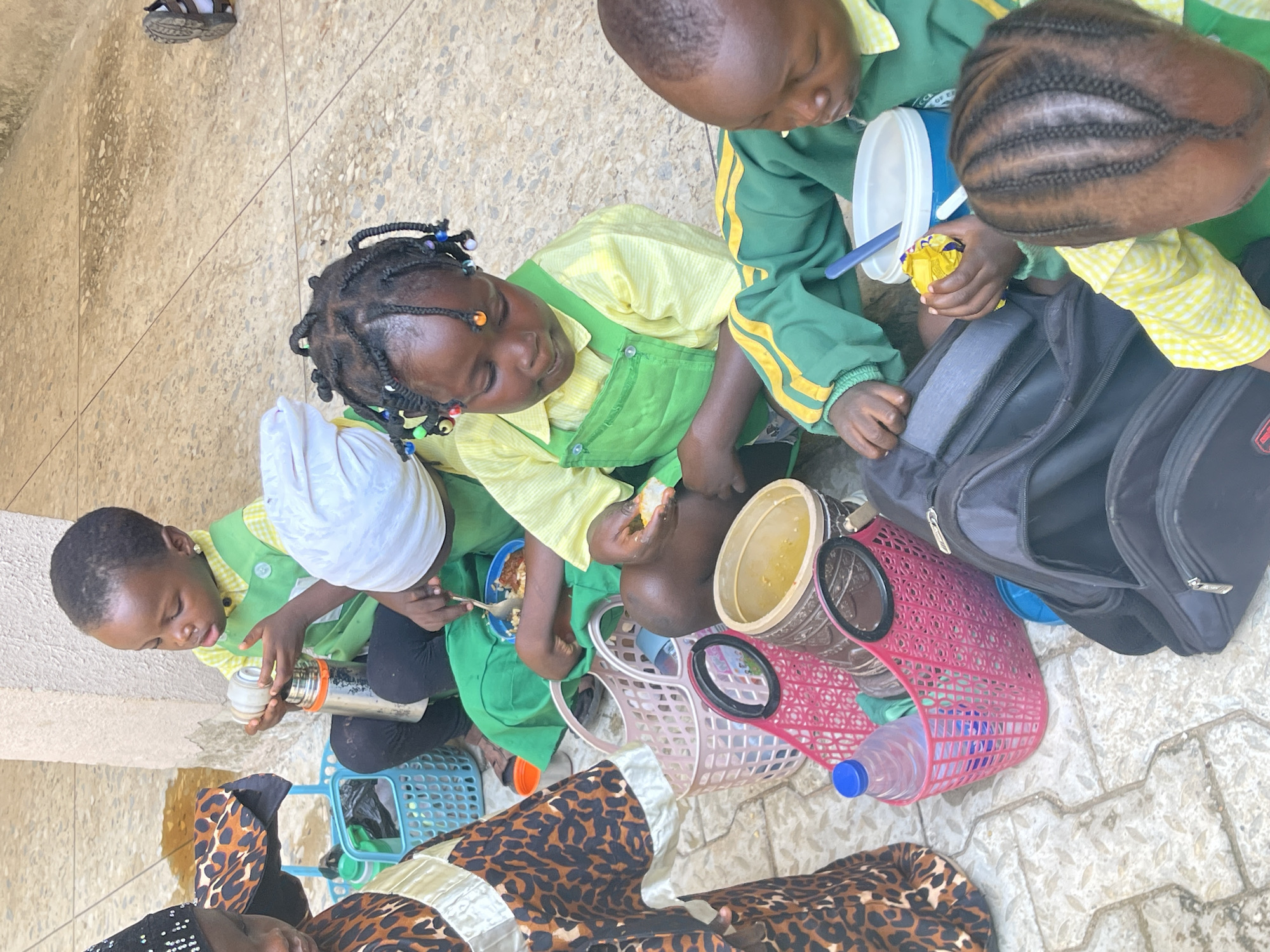 The image shows a group of children sitting on a paved surface, likely outdoors. They are eating from bowls and containers. A black backpack and several plastic baskets are nearby. The children are wearing school uniforms. The image suggests a mealtime scene in Akwanga, Nigeria.