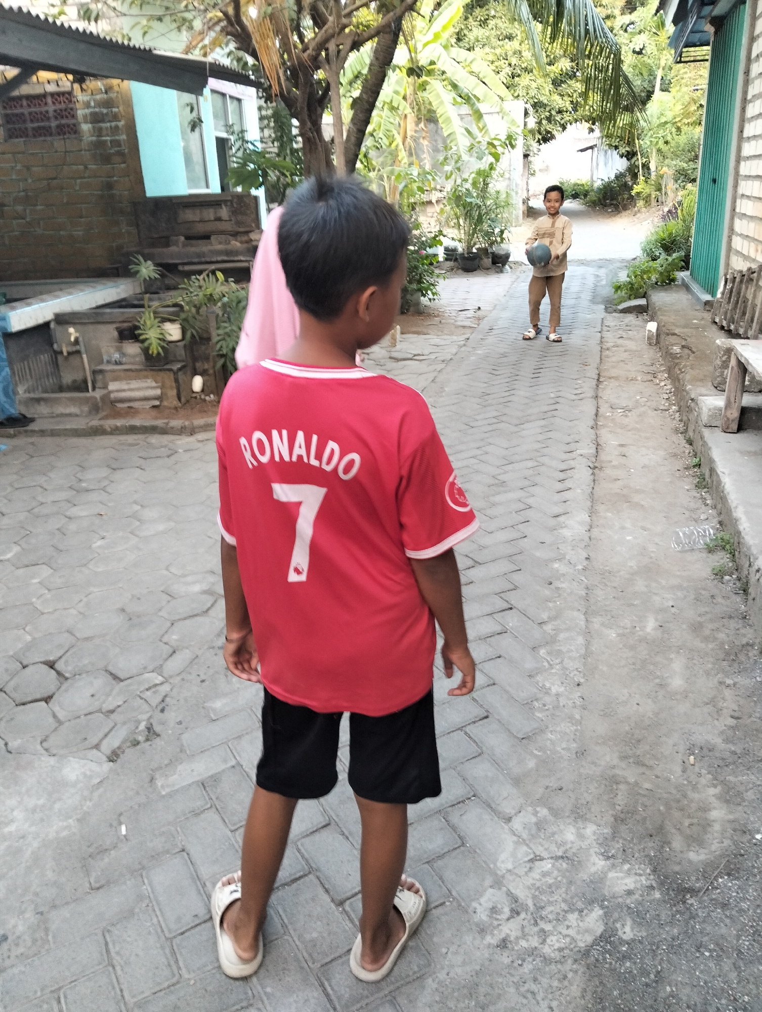 The image depicts two young boys in an outdoor urban environment, likely in Paciran, Indonesia, based on the context provided. In the foreground, a boy is seen from behind, wearing a red t-shirt with "RONALDO 7" printed on the back and black shorts. He is wearing white sandals. His stance suggests he is looking towards another individual.

In the middle ground, a second boy is facing the camera, holding a dark-colored ball. He is wearing a long-sleeved light brown shirt and brown pants, also with white sandals. The background reveals a pathway paved with interlocking concrete blocks, flanked by buildings and vegetation. One building has a brick facade and a blue section, with a corrugated metal roof. Lush green foliage, including palm trees, is visible throughout the scene, indicating a tropical or subtropical climate. The overall environment suggests a residential area or a public space within a town.