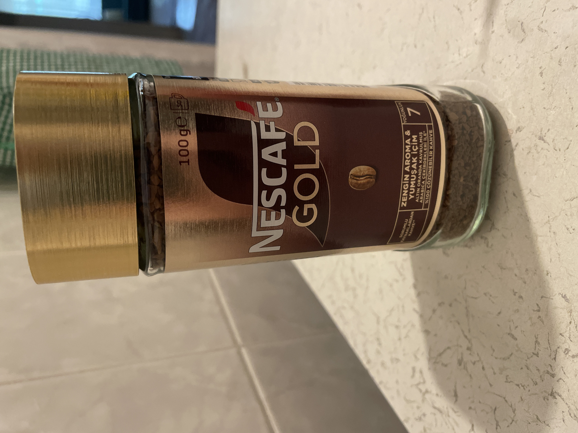 A close-up, eye-level shot shows a Nescafé Gold instant coffee jar. The jar is made of glass and is filled with coffee granules. The label is predominantly gold and brown, featuring the Nescafé Gold logo in white and gold lettering. Text on the label indicates "100 ge" and "ZENGİN AROMA & YUMUŞAK İÇİM" (Rich Aroma & Smooth Sip), along with "ALTIN ORANDA KAVRULMUŞ ARABICA ÇEKİRDEKLERİ İLE" (With Gold Roasted Arabica Beans) and "%100 ÇÖZÜNEBİLİR KAHVE" (100% Soluble Coffee). A small coffee bean graphic is visible below the main text. To the right, a number "7" is displayed within a red box, likely indicating intensity. The jar rests on a speckled white countertop, with a white tiled wall in the background. No people are present. This image is not identifiable as being specifically located in Kadıköy, Turkey, as there are no distinguishing landmarks or contextual clues.