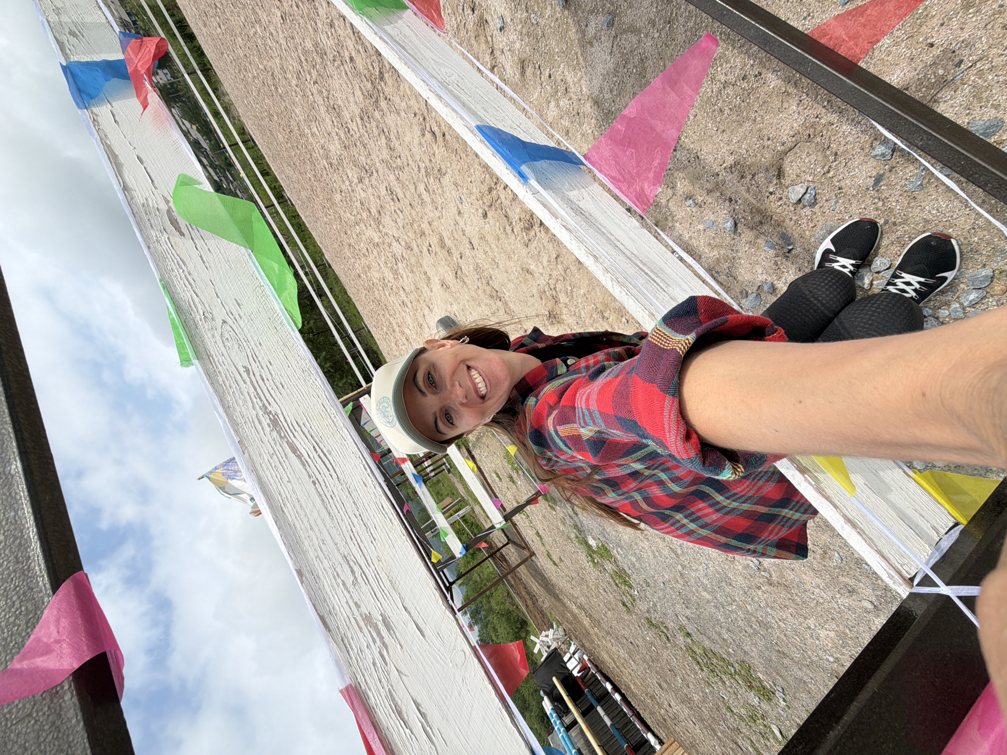 A woman in a red and black plaid shirt and a baseball cap is taking a selfie outdoors. She is smiling and her arm is extended as she holds the camera. She is wearing black leggings and black and white athletic shoes. The background appears to be a dirt or sand arena, possibly for equestrian events, with several white fences decorated with colorful triangular flags. In the distance, there are some trees and hills, suggesting a rural or semi-rural setting. The sky is overcast with some clouds. The image is taken from a slightly low angle, looking up at the woman. There is no indication of the city of Теплоозёрск, Russia in the image.