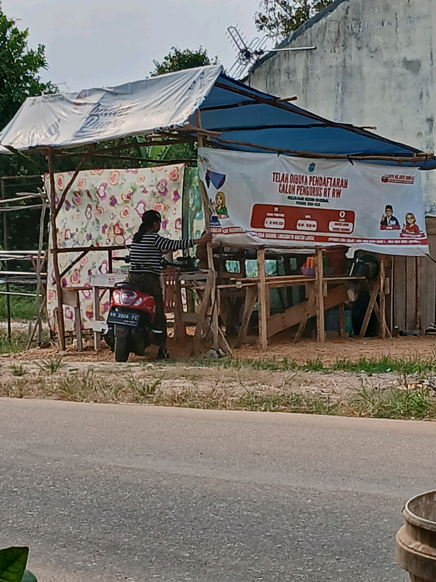 The image depicts an outdoor scene on a sunny day, likely in the city of Pangkalpinang, Indonesia, given the context provided. In the foreground, a paved road runs horizontally across the frame. To the left of the road, dry grass and sparse vegetation cover the ground.
Dominating the middle ground is a makeshift stall constructed from wooden poles and covered with a tarp. The tarp is predominantly white with some blue fabric underneath. A colorful, patterned cloth hangs from the front of the stall. A person wearing a black and white striped shirt and dark pants is standing next to a red scooter, which is parked on the ground in front of the stall. The person appears to be interacting with the stall, possibly a vendor.
A banner is prominently displayed above the stall, featuring text in Indonesian. The banner announces "TELAH DIBUKA PENDAFTARAN CALON PENGURUS RT RW" which translates to "REGISTRATION FOR RT RW CHAIRMAN CANDIDATES IS OPEN." Below this, there are graphics and some smaller text which is not clearly legible.
The background shows a weathered concrete wall and some trees. An old-fashioned television antenna is mounted on top of the wall. The overall impression is a humble, possibly rural or semi-urban setting, with a community-focused event being advertised.