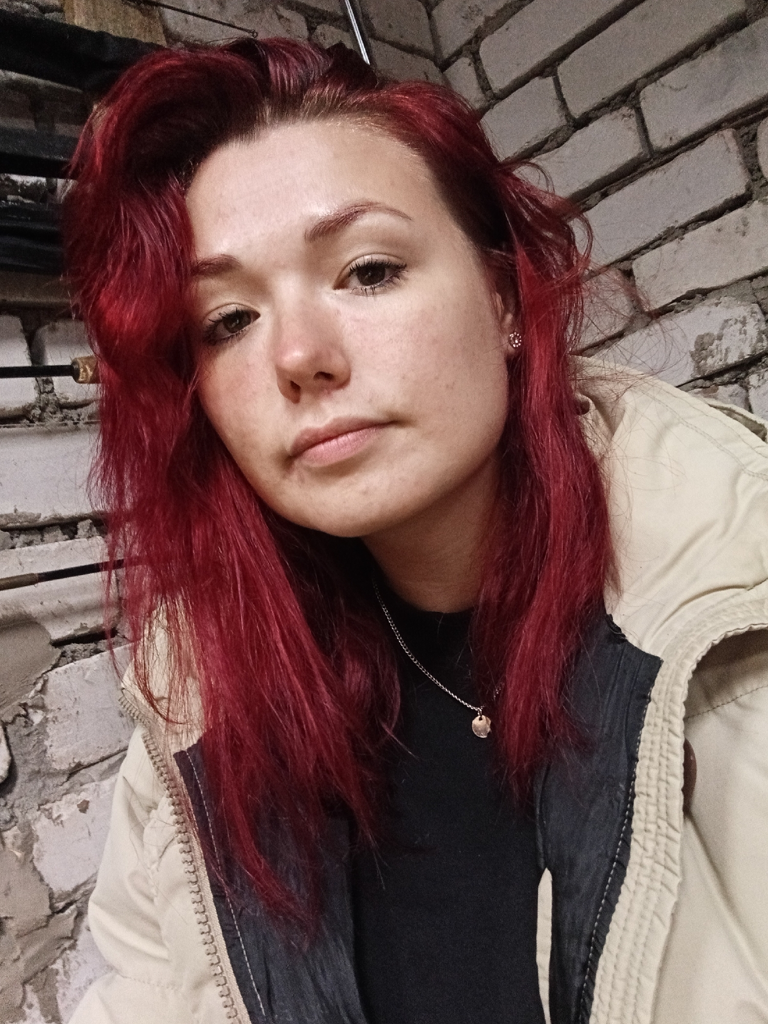 A young woman with vibrant red hair is pictured from the chest up, looking directly at the camera. She is wearing a light-colored jacket over a dark top, and a delicate silver necklace with a small pendant. Her face is illuminated by soft light, highlighting her features and a scattering of freckles across her nose and cheeks. The background consists of a textured brick wall, adding a touch of urban grit to the portrait. The overall impression is one of casual intimacy, as if captured in a candid moment.