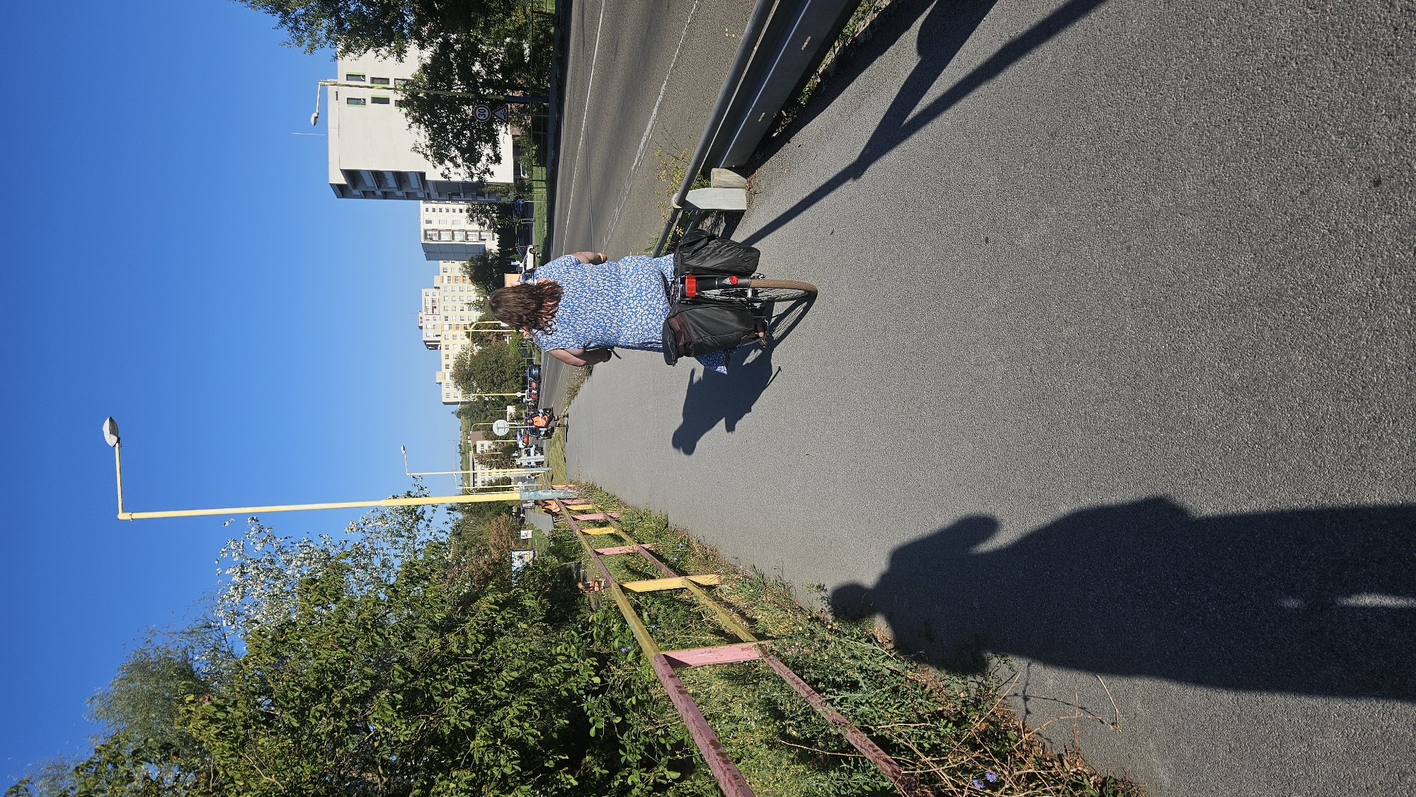 A person is cycling on a paved bike path alongside a road. The person is wearing a blue floral dress and has two black panniers on the back of their bicycle. The bike path is separated from the road by a metal guardrail. To the left of the bike path is a colorful railing, and beyond that, vegetation and trees. In the background, there are several apartment buildings and streetlights. The sky is clear and blue, indicating a sunny day. The scene is consistent with a residential area in the city of Prešov, Slovakia, possibly in a neighborhood like Sekčov which is known for its extensive green spaces and pedestrian/bike paths integrated with urban infrastructure.