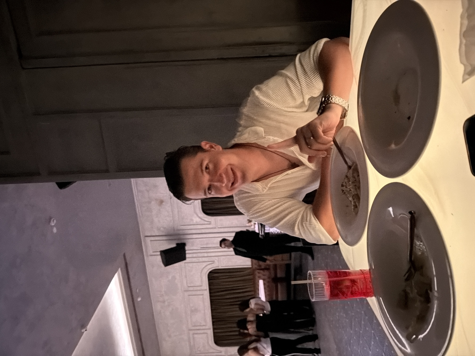 A young adult male, with dark hair and a light complexion, is seated at a brightly illuminated table, smiling directly at the viewer. He is wearing a light-colored, textured, collared shirt, a dark pendant necklace, a silver watch on his left wrist, and a ring on his right ring finger. His forearms rest on the tabletop. In front of him, a dark grey plate contains remnants of light-colored food, with a fork held by his right hand. To his immediate left, another dark grey plate holds a fork and appears largely empty, while a third empty dark grey plate is positioned to his right. A tall glass filled with a red beverage, ice, and a straw sits on the table to his left. The table is lit from underneath, casting an upward glow. In the mid-ground, a group of individuals, predominantly women in dark attire, are standing or moving towards the left. Behind them, a man in a dark suit stands looking left. The background reveals a dimly lit interior with ornate light-colored wall paneling featuring arched alcoves and dark curtains. A dark speaker is mounted high on the far wall. The right side of the frame shows a darker section of a paneled wall. The scene depicts an upscale restaurant or lounge located in New Delhi, India.