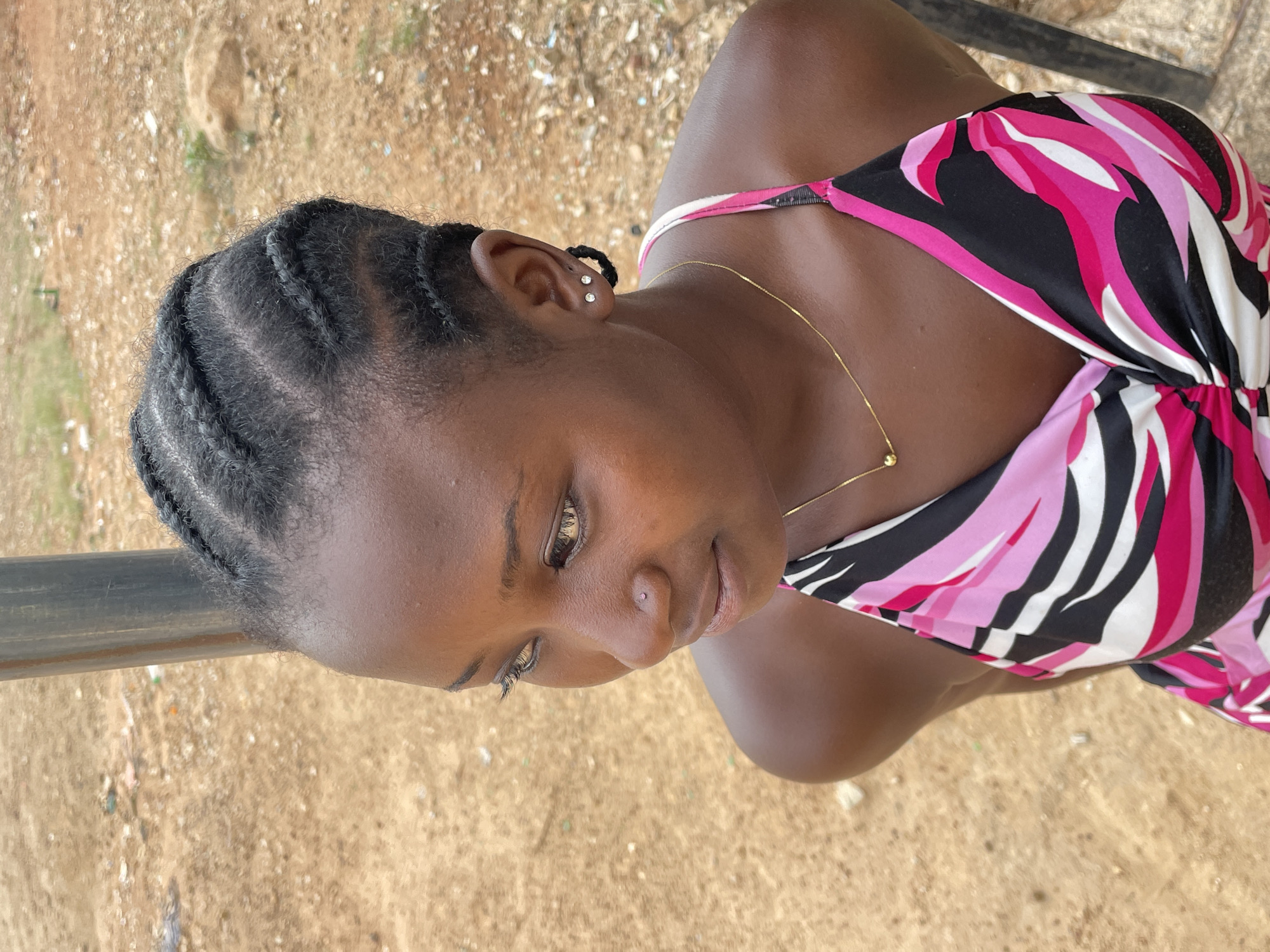A close-up portrait shows a young woman, likely from Barakin Akawo, Nigeria, looking downwards and to her right with a calm, contemplative expression. She has dark skin and her hair is styled in neat cornrows, braided tightly against her scalp. Her right ear is visible, adorned with two small, clear stud earrings. A tiny, clear nose piercing is also present on her nose. She is wearing a vibrant, patterned dress or top featuring a design of black, white, and various shades of pink. A delicate gold chain necklace with a small gold bead hangs around her neck. The background consists of dry, light brown ground, possibly dirt or sand, with scattered small debris. A dark, metallic bar runs horizontally across the upper left portion of the image. The scene is outdoors, well-lit, suggesting it is daytime. There is no visible text.
