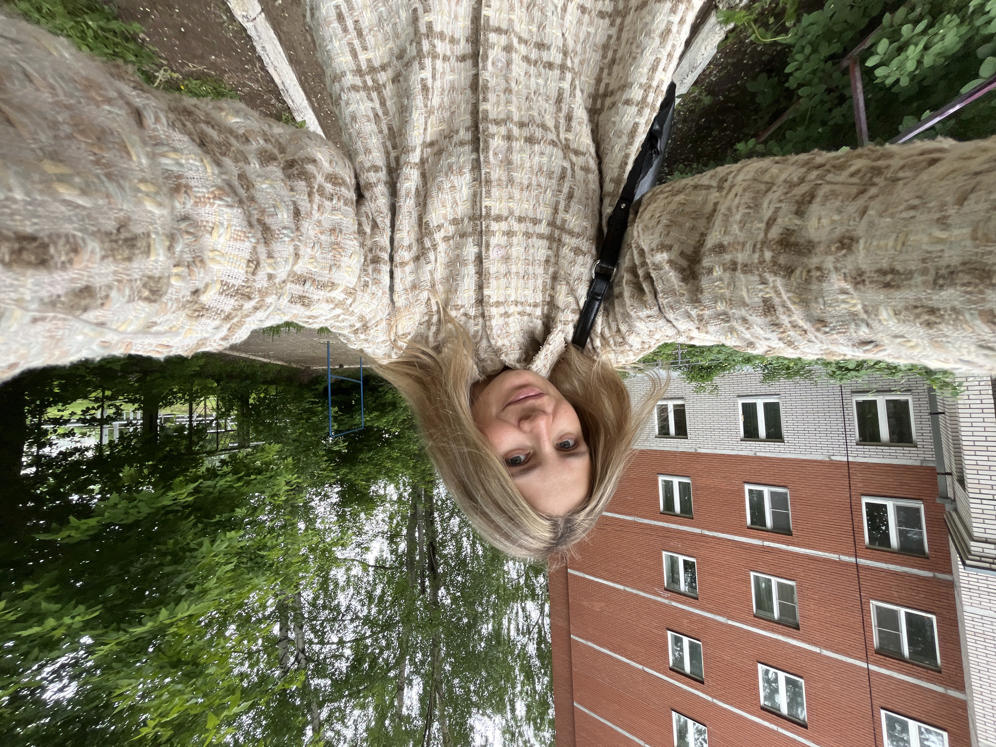 The image is a self-portrait taken outdoors, featuring a woman with blonde hair looking directly at the camera. She is seen from a low angle, holding the camera herself.
The main subject is a woman with long, straight blonde hair, which frames her face. She has light-colored eyes and a neutral, relaxed expression. She is wearing a textured jacket or shirt with a prominent beige and brown plaid pattern, made from a woven, possibly tweed-like, fabric. The garment has light-colored buttons down the front. A black strap, likely from a bag, is visible over her shoulder.
The setting is an outdoor urban or suburban environment. Behind the woman, to her right, is a multi-story building constructed of red brick, featuring light-colored horizontal bands and a lighter upper section. Numerous rectangular windows with white frames are visible across its facade. To her left, there are abundant green trees with dense foliage, indicating a park-like or heavily vegetated area. A paved or concrete ground surface is present near the base of the trees and building. Faint blue metal structures are partially visible among the greenery, possibly indicating playground equipment.
The activity depicted is the woman taking a selfie. The lighting suggests it is daytime, with soft, diffused light, possibly indicating an overcast sky or early morning/late afternoon. There are no harsh shadows. No text is visible in the image. The provided context places the location in Chaikovsky, Russia.