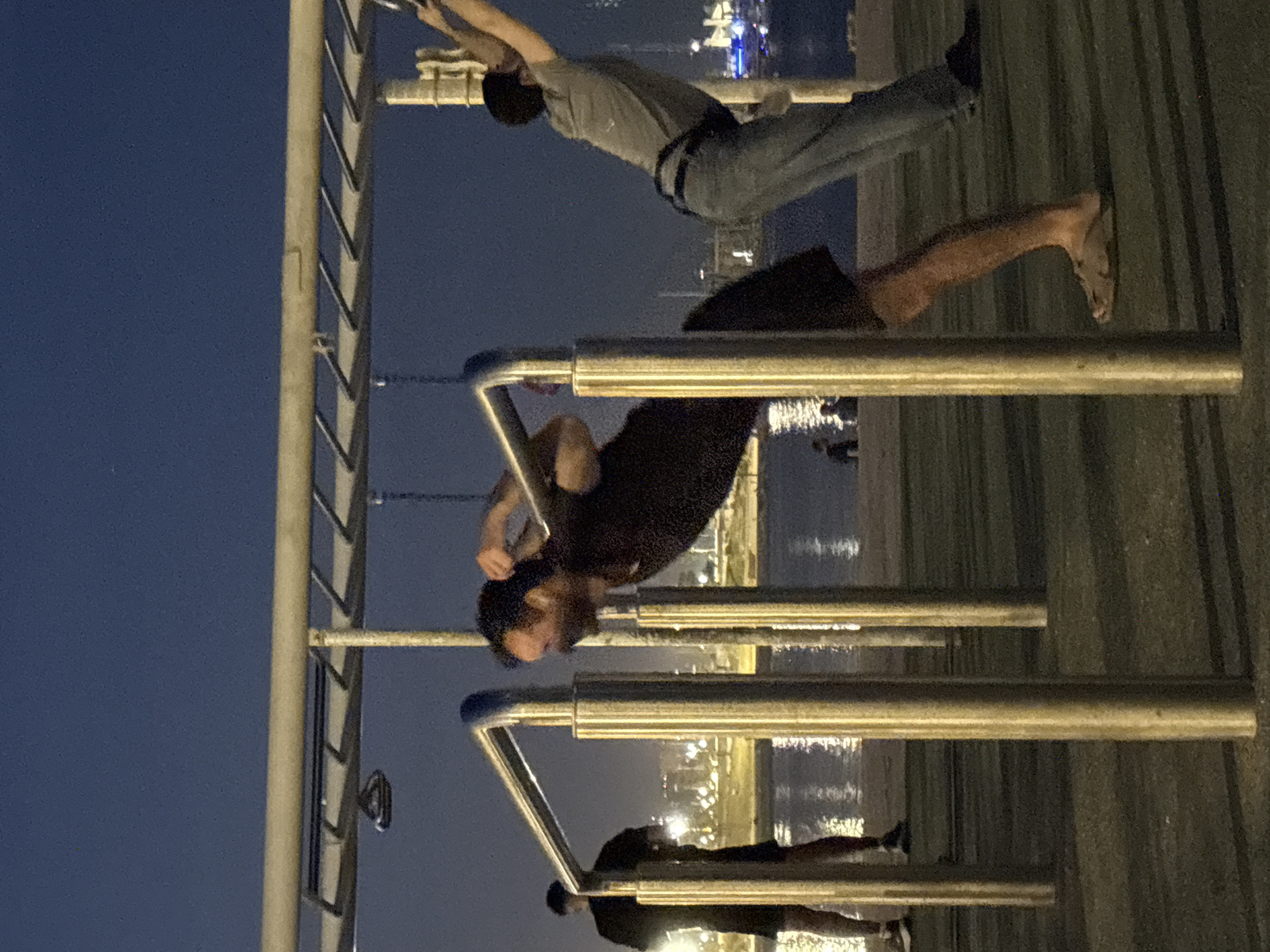 A nighttime image shows three individuals on parallel bars near a waterfront. The city lights in the distance suggest a possible location in Barcelona, Spain. The individual at the top is balancing, wearing a light-colored shirt, dark pants, and a dark belt. The individual in the middle is hanging from the bars, wearing a black shirt. The individual at the bottom is on the bars, wearing a dark-colored shirt and dark shorts. The foreground and background are blurry. The sky is dark blue.