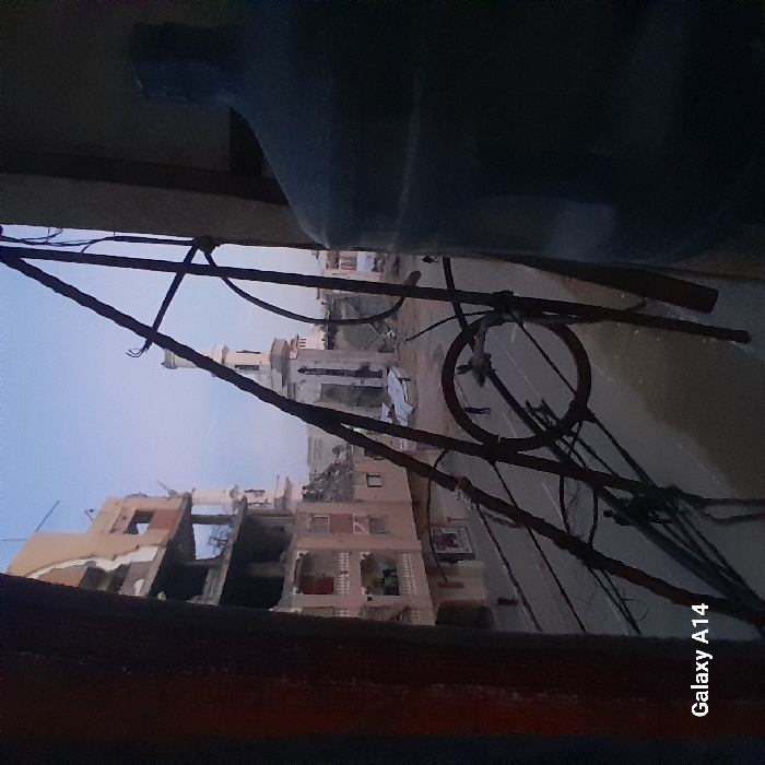 The image displays an outdoor urban scene, captured with a slightly askew perspective, as if looking out from a window or an elevated position. The overall lighting is soft and diffused, suggesting either an overcast day or early morning/late afternoon.
In the immediate foreground, to the left (if the image were rotated counter-clockwise), a large, dark blue or grey plastic container, possibly a water bottle, is partially visible, occupying the upper left quadrant. Below this, a dark, thick wooden frame or ledge forms the bottom border of the view, suggesting the vantage point is from inside a structure looking out.
The mid-ground is dominated by a complex network of dark, thin wires and possibly rebar, crisscrossing haphazardly across the frame. These wires obscure parts of the background, creating a cluttered and somewhat neglected impression.
Beyond the wires, the background reveals a street or unpaved road, which curves through the scene. On either side of this road, several multi-story buildings are visible. Many of these buildings appear to be in a state of disrepair or partial demolition, with exposed concrete, broken walls, and visible internal structures, suggesting damage or ongoing construction/demolition. One building in the distance, towards the center-right, stands out with a distinctive architectural feature resembling a mosque's minaret or a decorative tower, characterized by its lighter color and classical elements.
The sky is a pale, uniform blue-grey, indicating a lack of direct sunlight or an overcast sky. No people are visible in the scene.
The only discernible text in the image is a watermark in the bottom right corner, which reads "Galaxy A14", indicating the type of device used to capture the photograph.