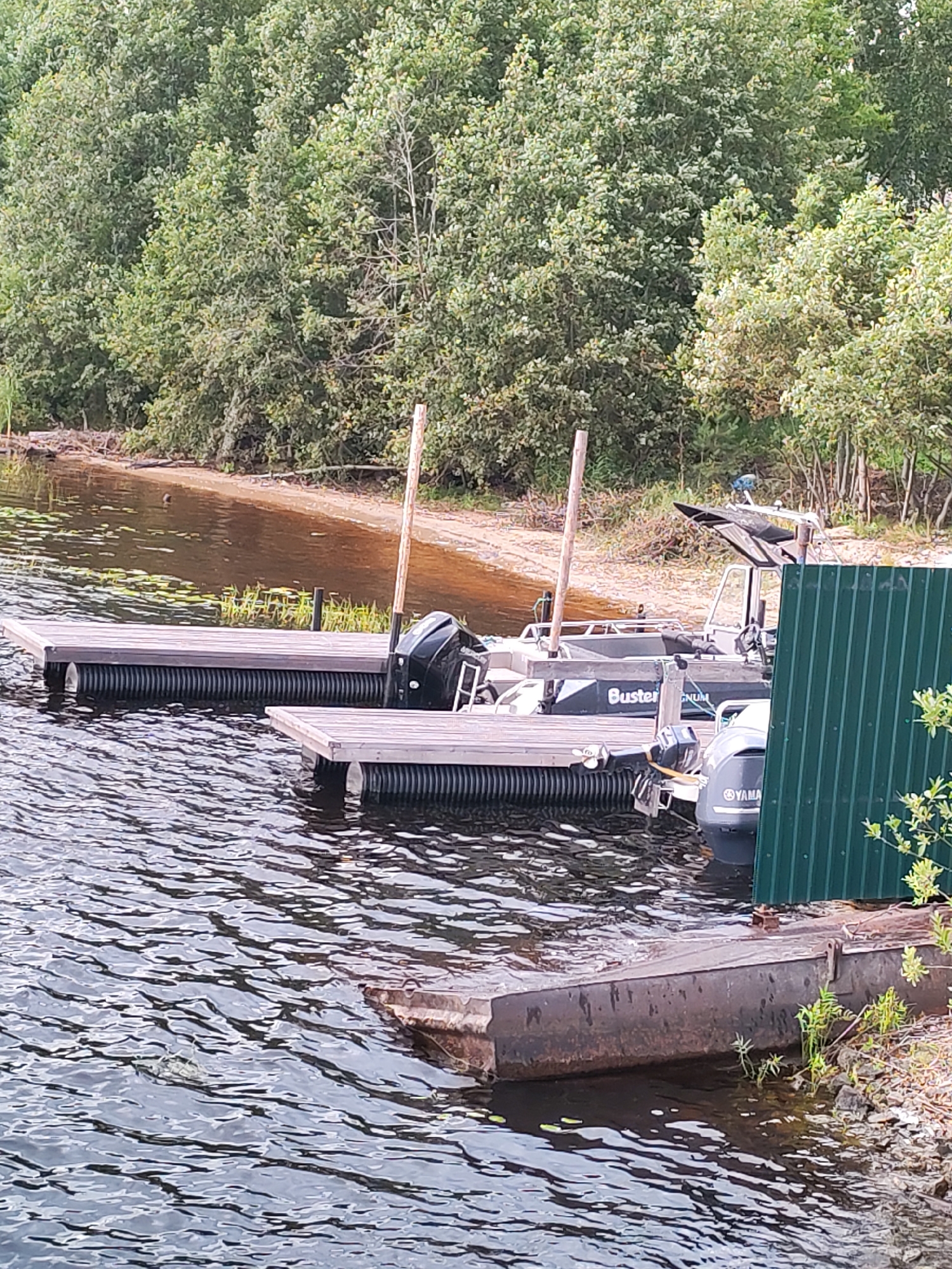 A scene on the water's edge in Shalsky, Russia, features two wooden docks with black, ribbed flotation devices underneath, moored alongside a sandy shoreline with dense green trees in the background. Two boats are visible. The boat closest to the viewer has a black outboard motor and a dark hull with "Buster Magnum" written on its side. A second boat, partially obscured by a green metal fence, has a gray outboard motor with a Yamaha logo. The water is choppy, reflecting the overcast sky. The shoreline is covered in sand and scattered with fallen branches.
