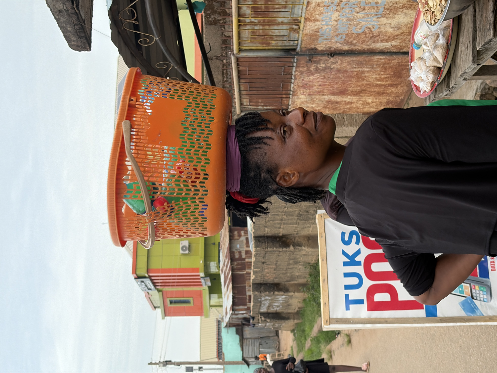 A woman with braided hair and a purple headband is carrying an orange basket on her head. She is wearing a black top with a green neckline. To her left, there's a sign reading "TUKS POINT" with an image of a mobile payment device and a blue credit card. Above her head is a metal tray containing small white bags and nuts. In the background, there's a street scene with various buildings, including one painted green and red. The scene is taking place in Jos, Nigeria. In the far background are power poles, and a person walking on the ground. The sky is overcast.