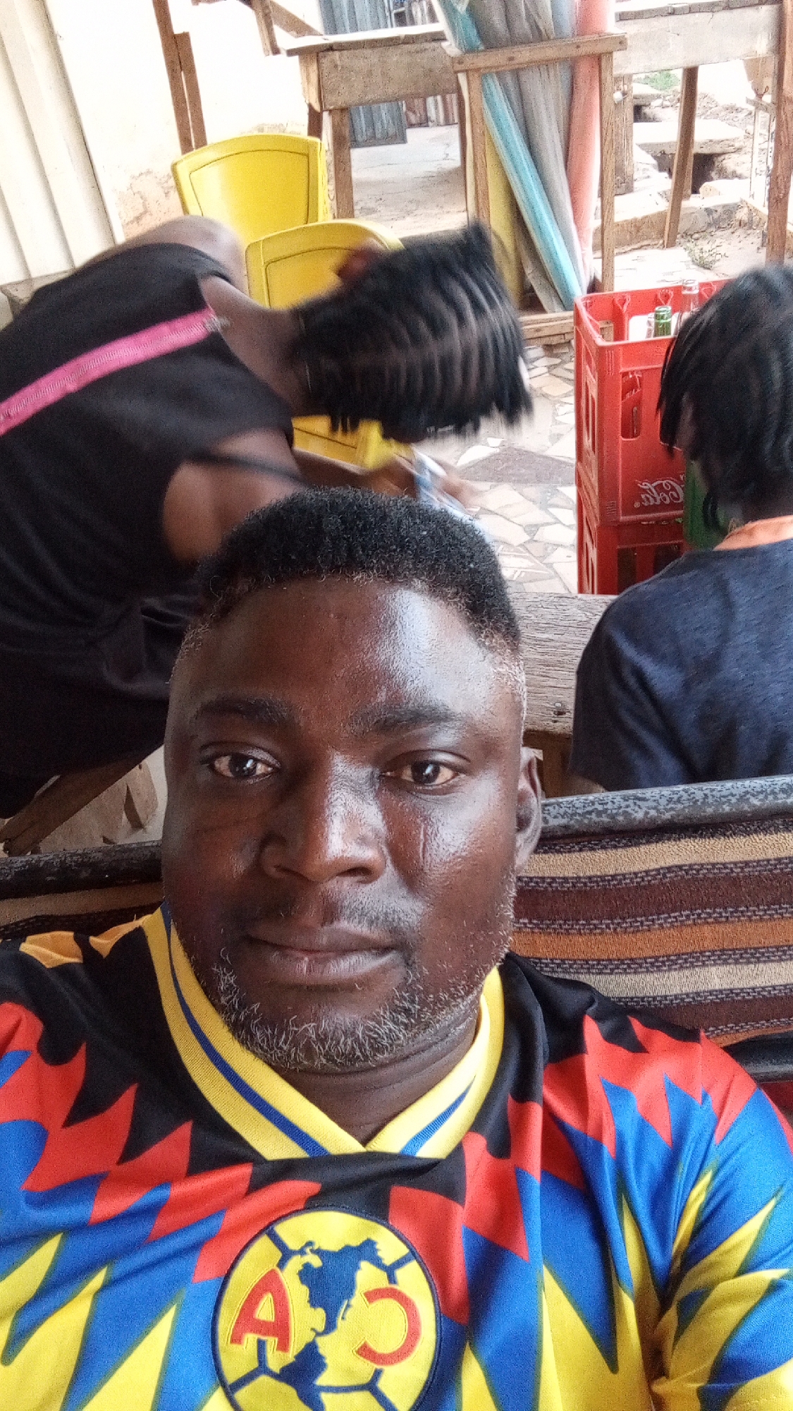 A dark-skinned man takes a selfie in what appears to be an outdoor or semi-outdoor setting in Sabon Tasha, Nigeria. He is positioned in the foreground, looking directly at the camera with a neutral expression. He has short dark hair with faded sides and a short, salt-and-pepper beard, and a small scar or mark is visible on his left cheek. He is wearing a vibrant soccer jersey patterned with black, red, blue, and yellow. The jersey prominently features a circular logo on the chest, depicting a globe outline with the letters "A" and "C", identifying it as a Club América design.
In the background, two other individuals are partially visible. To the left, a person with dark braided hair wears a black shirt with a pink strap across their back. To the right, another person is seen from behind, wearing a dark shirt and dreadlocks. Behind these figures, the setting includes rustic wooden tables and structures, several bright yellow plastic chairs, and a red plastic crate containing bottles. The "Coca-Cola" logo is faintly visible on the side of the crate. The ground is a mix of dirt and possibly paving, with corrugated metal sheets visible in the distant background. Some light-colored and pastel fabric strips hang between wooden supports. The overall scene suggests a casual, informal gathering place, likely during daylight hours under shaded conditions.