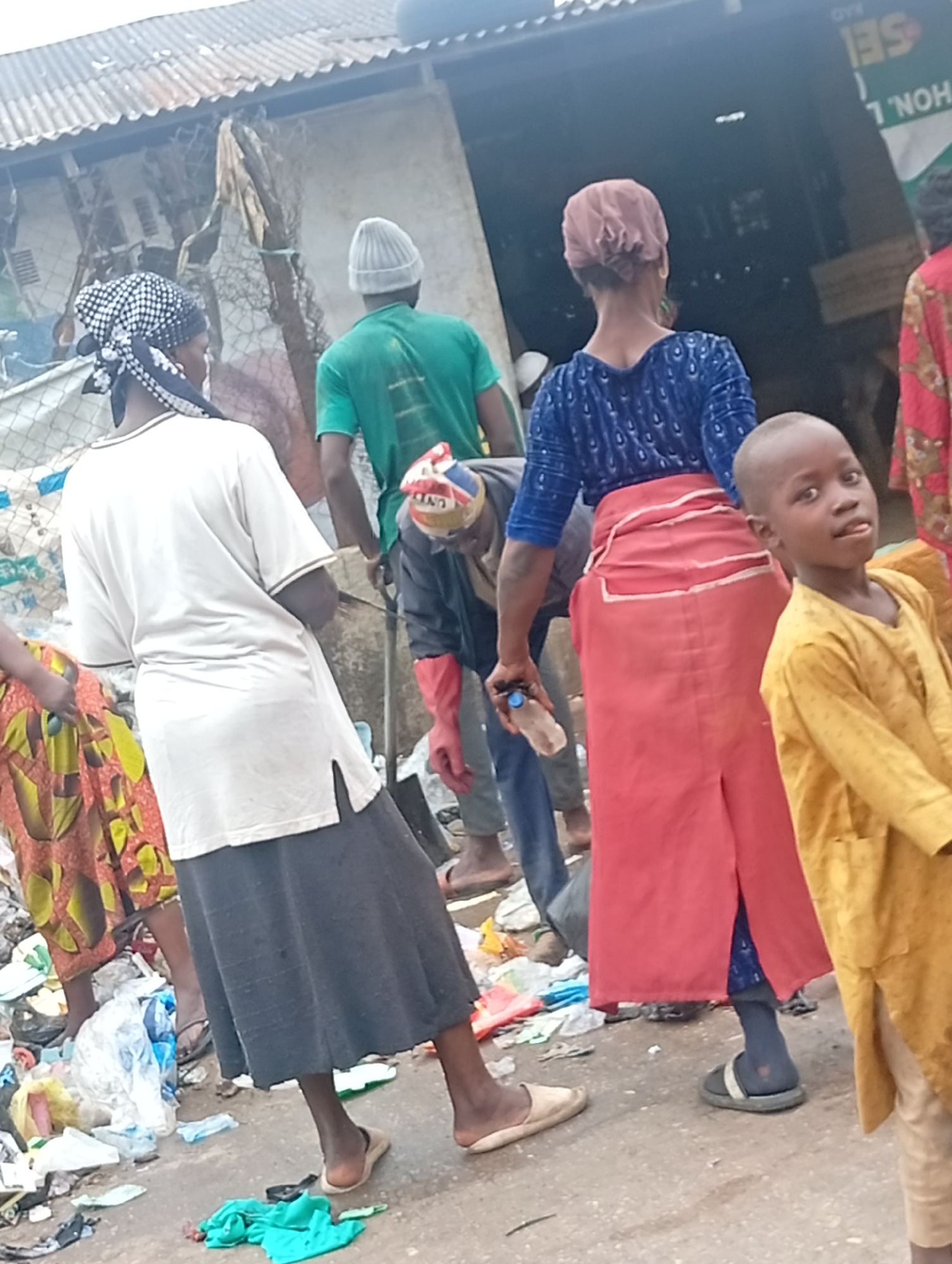 The image shows a street scene in what appears to be Maiburiji, Nigeria, based on cultural and environmental factors. Several individuals are present, including adults and a child in the foreground wearing a yellow garment. The adults are dressed in a mix of traditional and casual attire, with a woman in a blue top and red skirt being prominent. One person wears a green shirt and another a white shirt and black skirt. Many individuals wear head coverings. A man in a gray hooded jacket is holding a water bottle.
The environment is visibly littered with garbage and discarded items, suggesting a lack of waste management. Buildings in the background are constructed with corrugated metal and other rudimentary materials. The scene suggests a low-income area. The interactions of the individuals are not overtly clear.