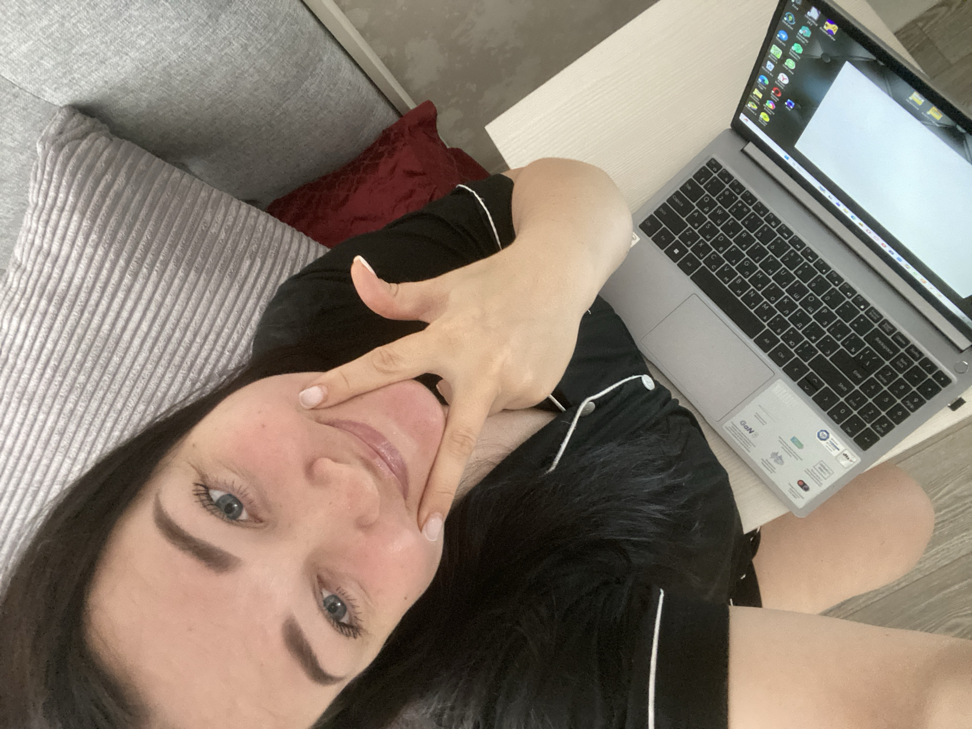 A person, likely female, is taking a selfie from an overhead perspective while lying down indoors. She has dark hair, blue eyes, and is looking directly at the camera with a gentle smile. Her right hand is raised, with fingers forming a "peace sign" or "victory sign" near her face. She has light-colored nail polish on her fingernails.
She is dressed in black loungewear or pajamas with white piping. Her head is resting on a grey, ribbed pillow, which is on a grey sofa. A dark red cushion is also visible behind her. To her right, a silver-grey laptop with a dark keyboard is open on a light-colored side table. The laptop screen shows a desktop with numerous application icons on the left side and a large white rectangle, possibly an open document or browser window, dominating the rest of the display. Visible text on the laptop keyboard includes Latin and Cyrillic characters. Stickers near the touchpad include "GaN" and other certification logos. Her bare legs are visible in the lower right portion of the frame, resting casually. The overall setting appears relaxed and domestic.