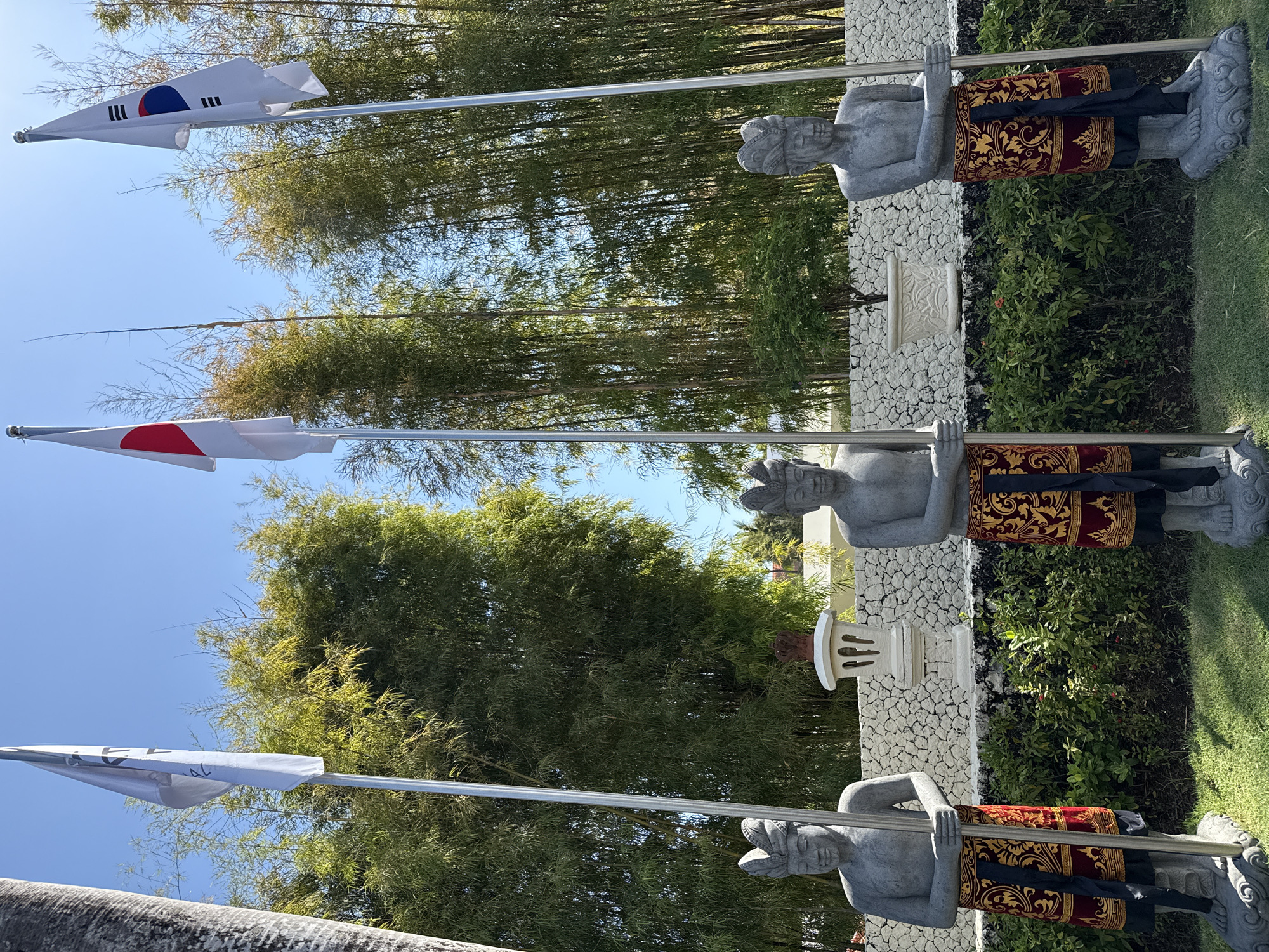 A brightly lit outdoor scene features three traditional Balinese-style stone statues holding flagpoles, set against a backdrop of lush bamboo trees and a clear blue sky. The grey stone statues, resembling guards or gatekeepers, stand in a row. Each is adorned with intricate black, red, and gold patterned garments, typical of Balinese *saput* and *kamen*. Their feet are also intricately carved.
From left to right, the flags displayed are the national flag of South Korea (Taegeukgi), the national flag of Japan (Hinomaru), and a white flag with black text that appears to read "BALI." The flags are gently waving, suggesting a light breeze.
Behind the statues, a low white stone wall with a textured surface separates the display from a dense grove of tall green bamboo. Between the second and third statues, an ornate white stone pedestal or small shrine is visible behind the wall. The foreground and surrounding area are covered in neatly trimmed green grass and low-growing vegetation.
The clear blue sky and strong sunlight indicate midday or early afternoon on a sunny day. The artistic style of the statues and the tropical flora firmly place the scene in Bali, Indonesia, consistent with the Kuta Selatan location context. The display of flags suggests a welcoming gesture or an international theme, possibly at a resort or cultural venue.