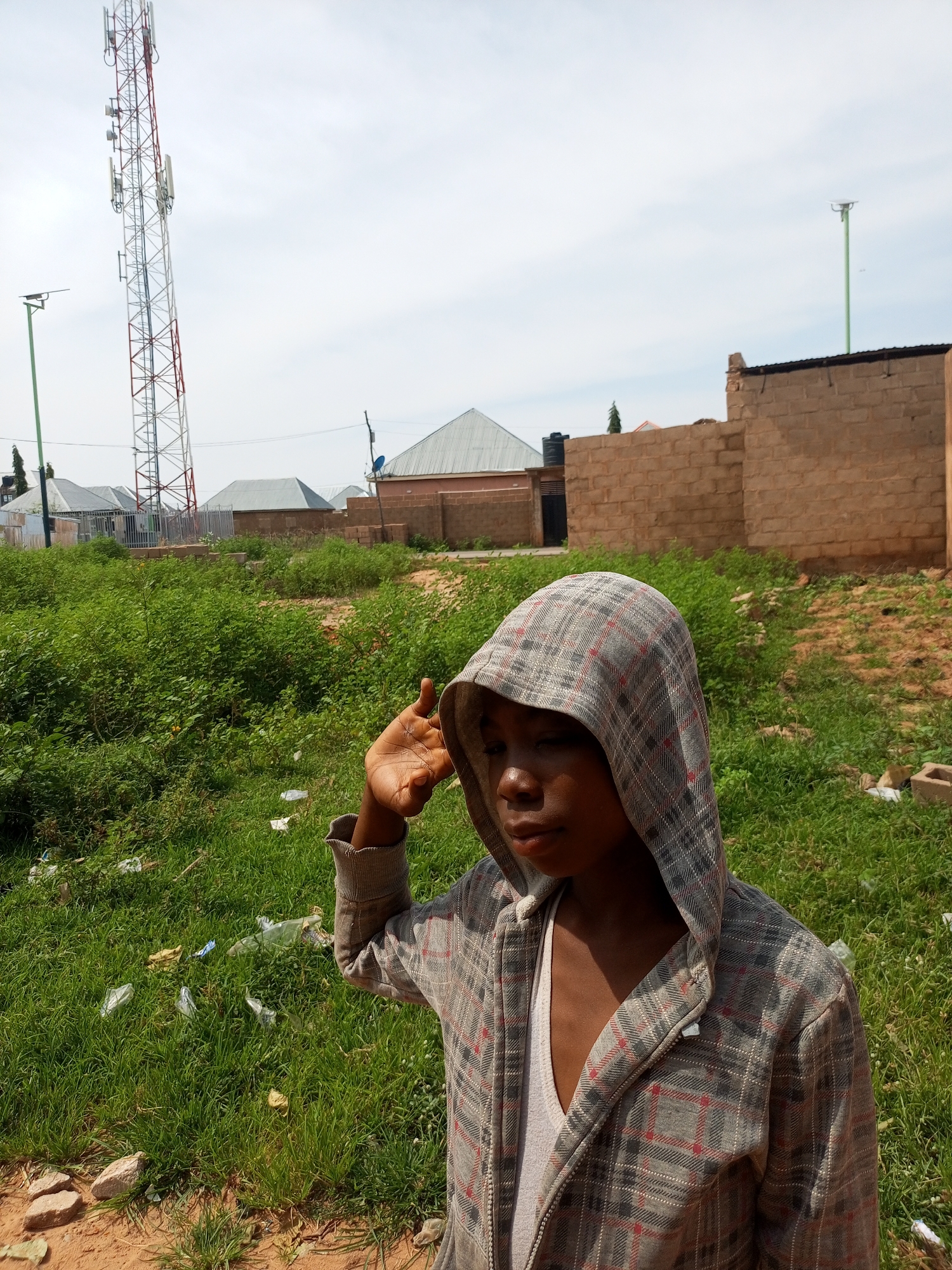 The image shows a young person wearing a plaid hooded jacket and a white v-neck shirt. The person is standing in an outdoor environment, with a grassy area containing litter in the foreground. In the background, there are buildings, a tall communication tower, and a cloudy sky. The scene appears to be in a semi-urban or rural area. The specific location in the city of Tempure, Nigeria, cannot be determined from the visual information alone.