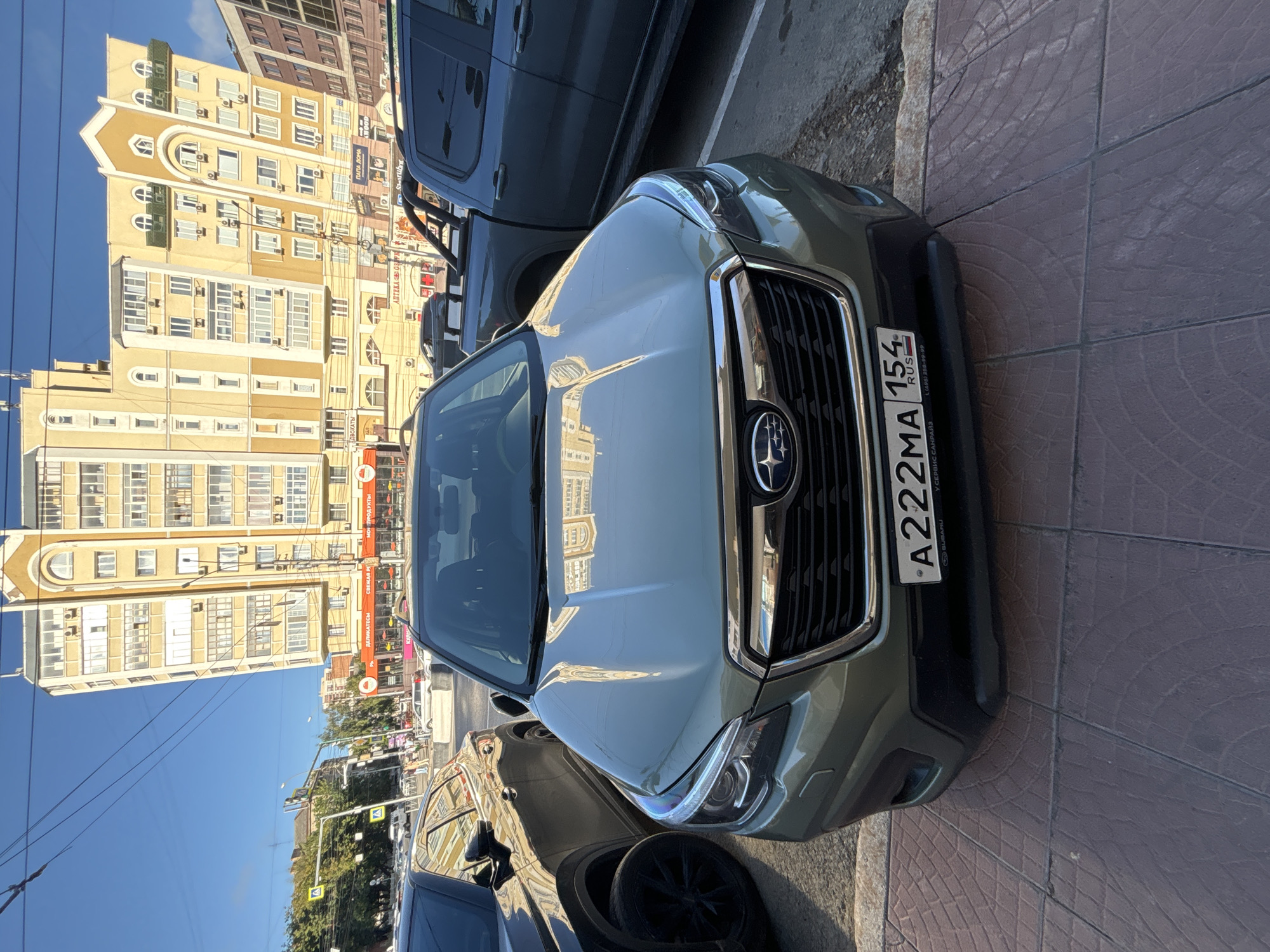 In Novosibirsk, Russia, a green Subaru Forester is parked on a tiled sidewalk in front of a multi-story building. The car's license plate reads "A222MA 154 RUS." To the left of the Subaru, a dark-colored SUV is parked, and to the right, a dark-colored pickup truck is visible. The building behind the cars features a cream-colored facade with numerous windows and balconies, and appears to house commercial establishments on its ground floor, indicated by visible signage. The sky is clear and blue, suggesting a sunny day. The scene depicts a typical urban street environment.