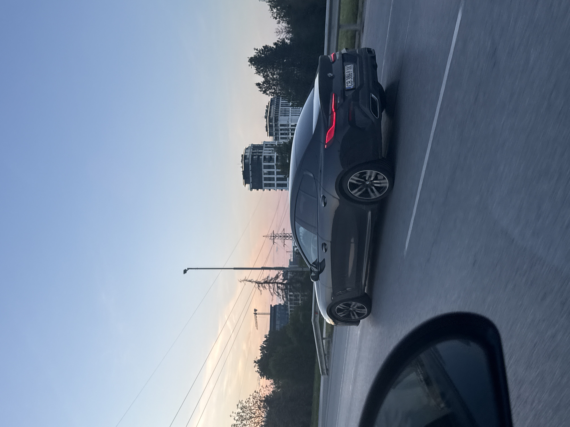 The image depicts a dark gray BMW car with a license plate number “CB 3869 XK” driving on a multi-lane road in Sofia, Bulgaria. The photograph was presumably taken from a car in an adjacent lane as the passenger side mirror of the photographer’s car is visible in the bottom of the frame. Buildings and infrastructure elements are visible in the background including apartments, powerlines, a construction crane, and streetlights. The sky shows a sunset with hues of orange and pink.