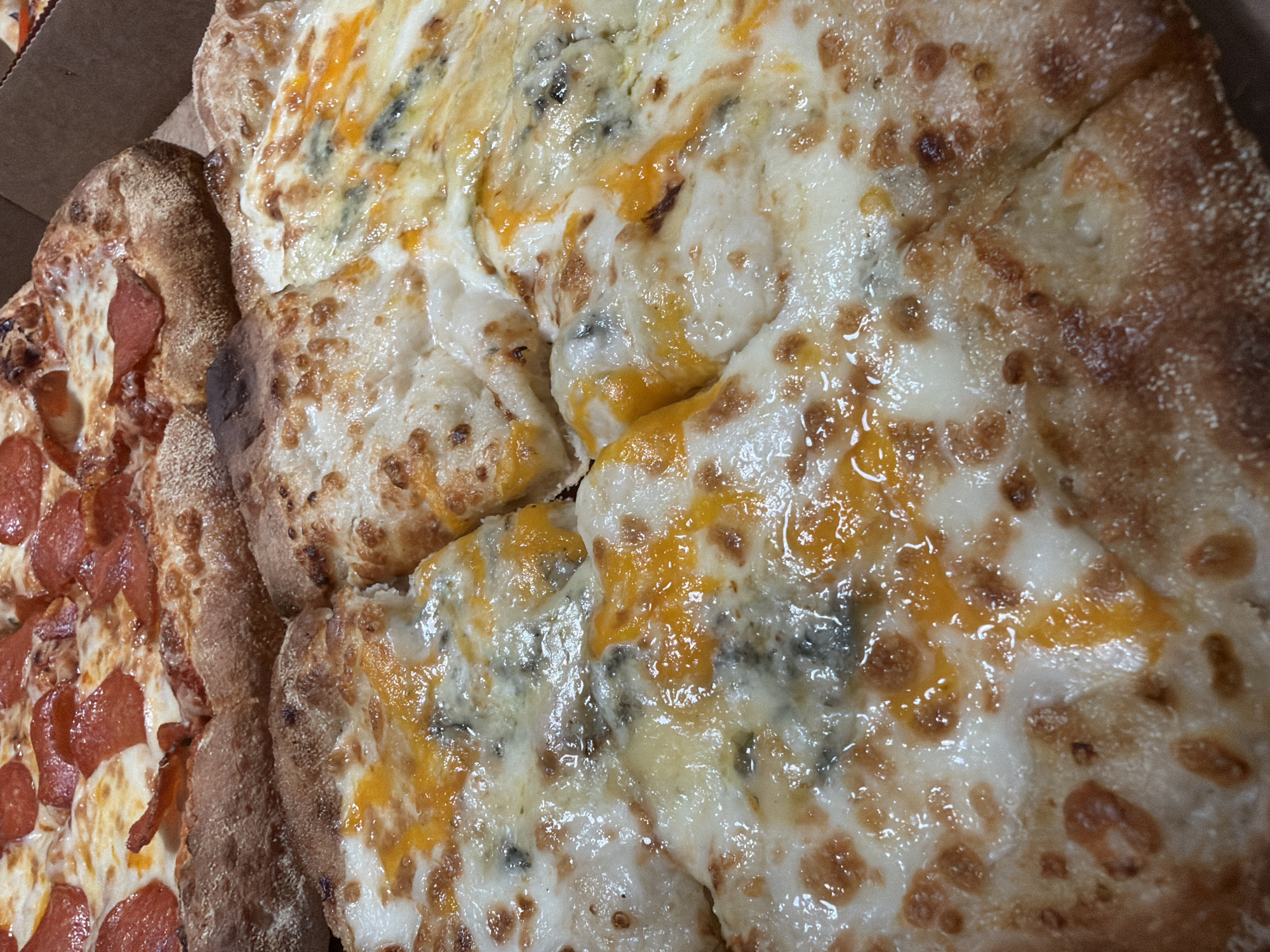 The image shows two pizzas in a cardboard box, likely taken from a high angle. On the left is a pizza with pepperoni. The right pizza is covered in cheese, with some areas showing yellow and bluish spots. The crust is bubbly and slightly browned. The pizzas are sliced into pieces. There is no information provided in the image to locate the scene in Votkinsk, Russia, or any details about people, actions, or interactions.