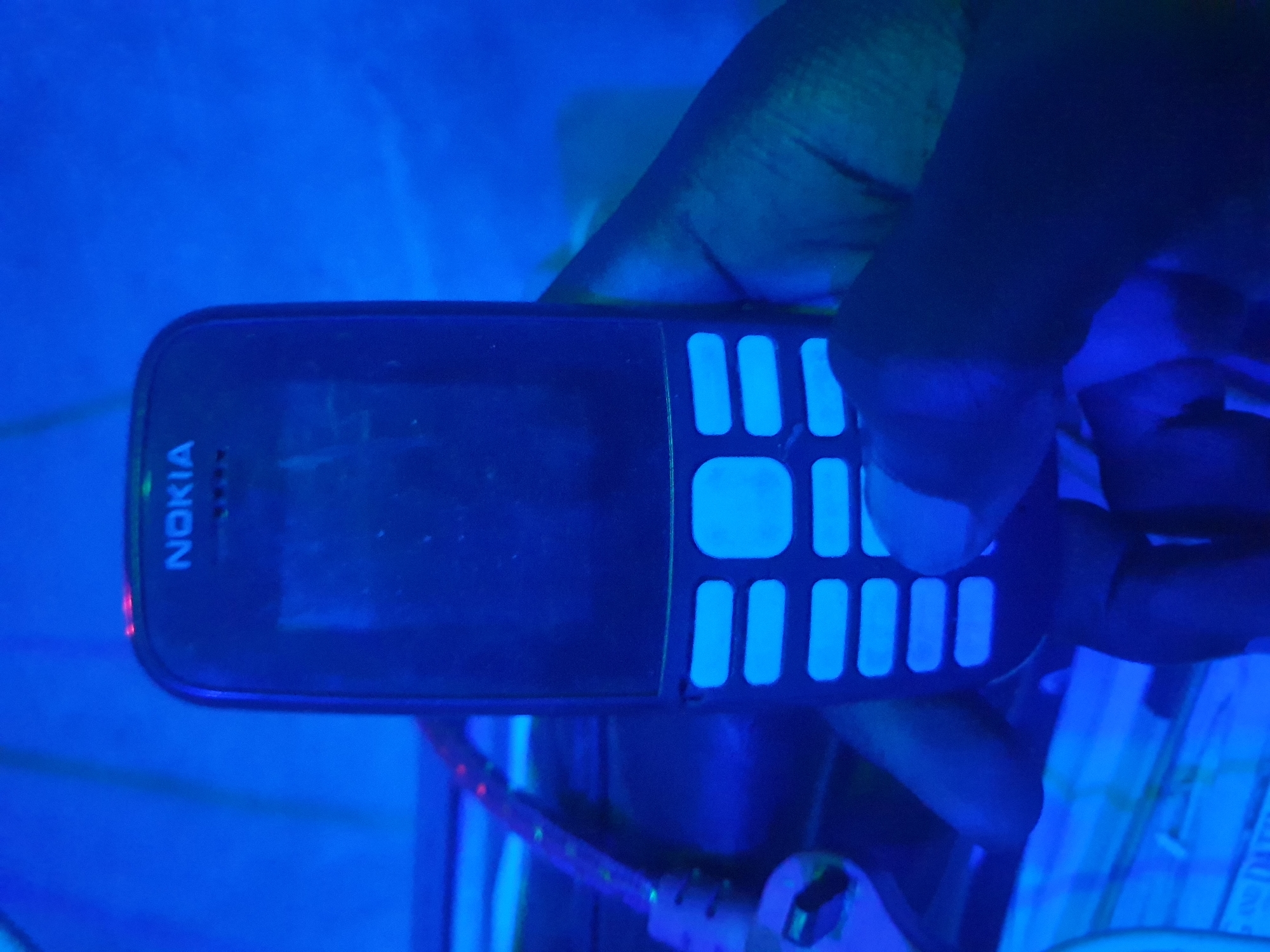 A dark-skinned hand holds a black Nokia feature phone under blue lighting. The phone's screen is dark, and the keypad is illuminated with bright blue light. The background is a blurred blue and green environment, possibly an indoor setting in Anilelerin, Nigeria, with some wires and electronics visible. The lighting creates a stark, high-contrast effect.
