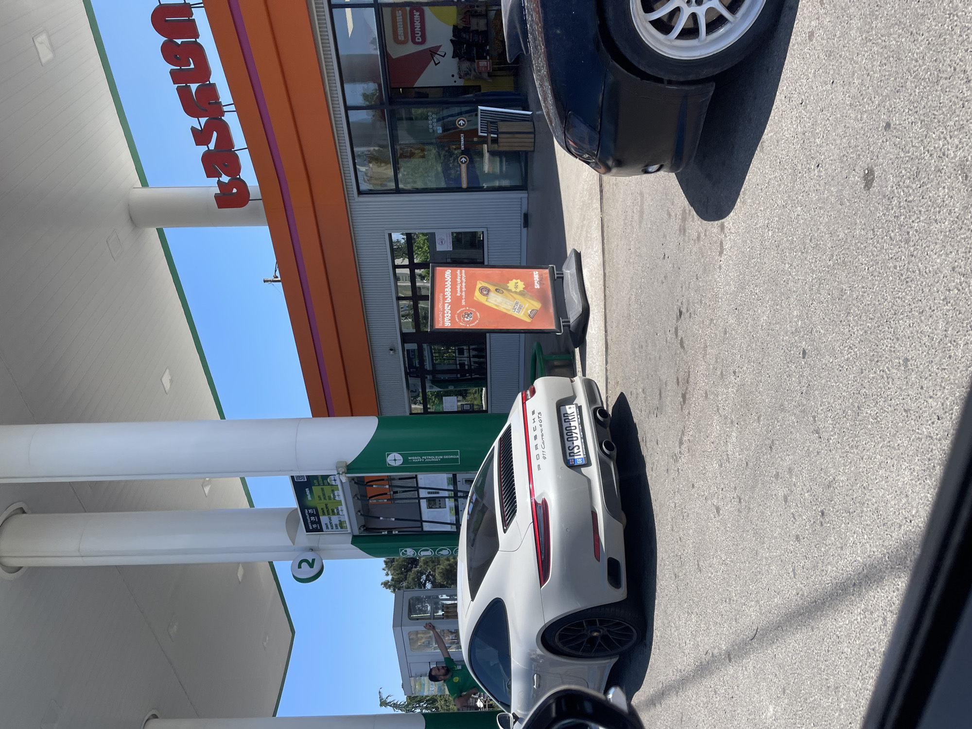 The image captures a light gray Porsche 911 Carrera 4 GTS (Georgian license plate GE RS-090-RR) positioned at pump number 2 of a Gulf gas station under a clear blue sky. The gas station's canopy is white with a green trim, supported by white cylindrical pillars. Pump 2 displays fuel types and prices in Georgian, including designations for "-15, -20, -25, Super, Premium, Euro Regular, and Diesel." A male gas station attendant, wearing a green uniform shirt, is visible to the left of the Porsche, leaning against a gray utility box.
The adjacent gas station convenience store features an orange facade with a prominent purple horizontal stripe. Red Georgian script, spelling "გალფ" (Gulf), is mounted on the orange facade. The lower section of the building consists of large glass windows and horizontal ribbed metal paneling. Inside the store, a "DUNKIN'" logo is visible through the windows, indicating a Dunkin' Donuts outlet. An "ENTRANCE" sign with an arrow is affixed to the store's glass door. An A-frame sign stands on the pavement near the entrance, displaying an advertisement in Georgian for a "Spark" product, which appears to be a tobacco item, with a "-50%" discount.
In the immediate foreground on the right, the front bumper and a white multi-spoke wheel of a black vehicle are partially visible. The ground is paved with asphalt. Green foliage from trees is discernible in the background. The scene is brightly lit by direct sunlight, casting distinct shadows. The presence of Georgian script on signage and the Georgian license plate ("GE") confirms the location as Tbilisi, Georgia, where Gulf is a recognized gas station chain.