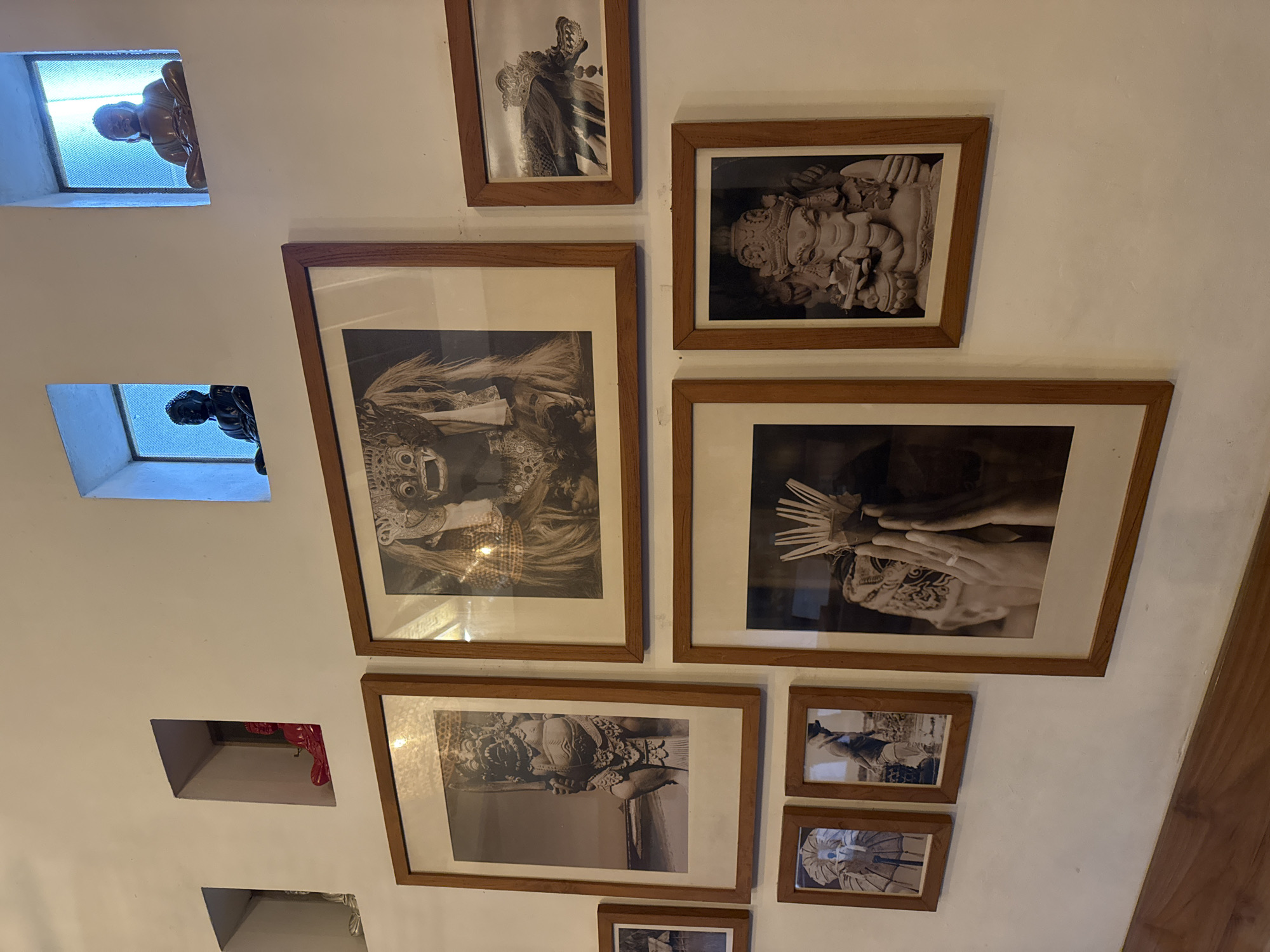 The image displays a white wall adorned with an artful arrangement of black and white framed photographs and recessed niches, indicative of an interior setting, likely in Kuta Selatan, Indonesia.
Seven photographs, all framed in natural wood, are mounted on the wall. The subjects within these images are primarily cultural or traditional. A large frame on the middle left features a figure in elaborate ceremonial attire, possibly a Balinese dancer or masked performer. Above it, a smaller frame shows another figure in ornate dress. To the right, a prominent photograph depicts a sculpted head, resembling Ganesha or another deity. Adjacent and below, a large frame captures hands engaged in what appears to be a ritualistic act, holding fan-like objects. Below the central left frame, another large photograph showcases an intricately carved deity or statue. Two smaller frames in the bottom right corner display less distinct cultural motifs.
The wall also incorporates several rectangular niches. The topmost niche contains a light-colored, serene Buddha statue, illuminated from behind by a bright, possibly blue-hued light. Below it, a similar but darker Buddha statue resides in another backlit niche. A lower niche reveals a glimpse of red fabric or an object, and a fourth niche is partially visible. The overall composition blends traditional art and sculpture, creating a culturally immersive visual display. The lighting suggests an indoor setting, with some backlighting accentuating the figures in the niches.