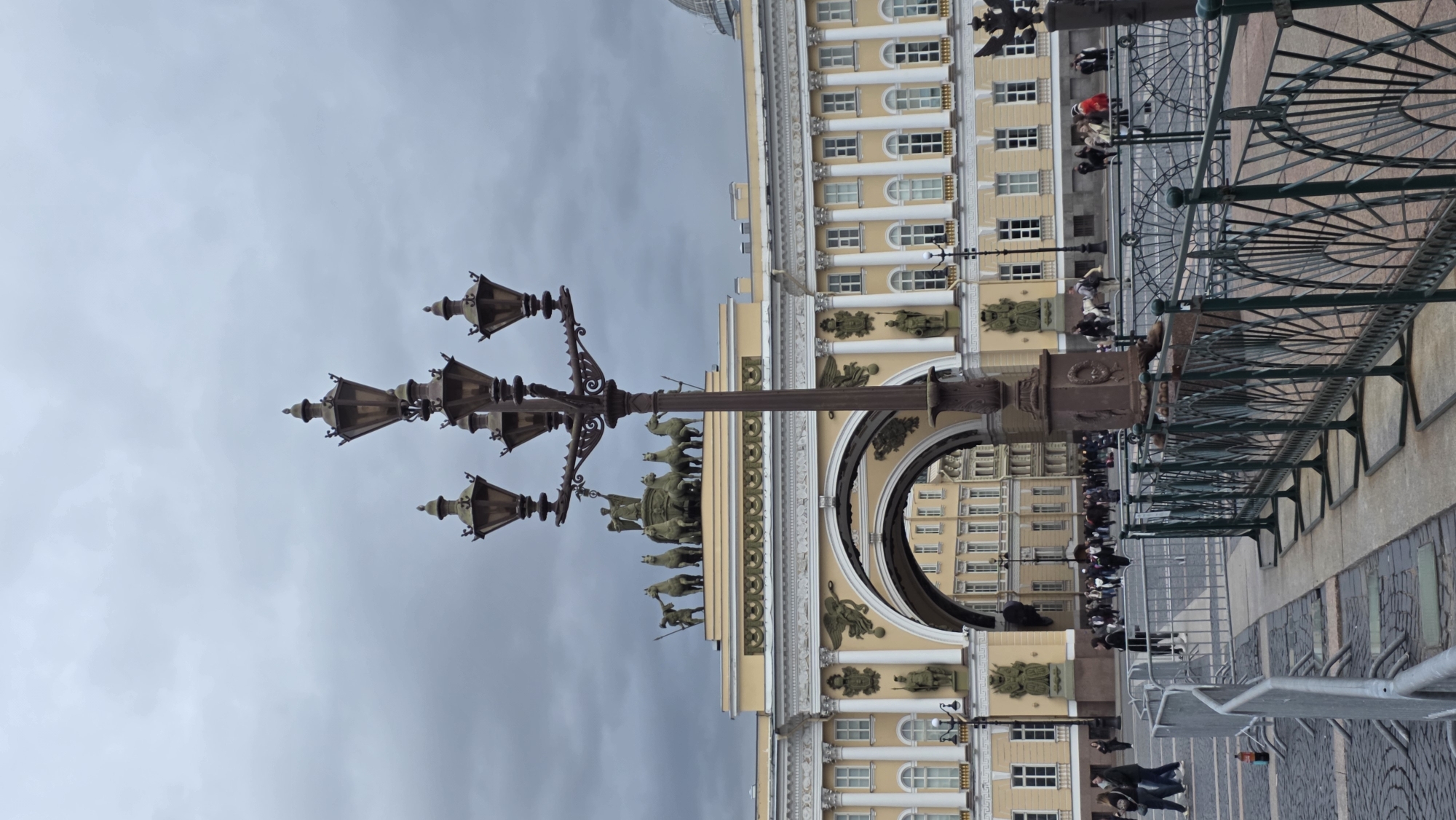 The image displays a vertical shot of a cityscape under an overcast sky. In the foreground, a detailed, dark-colored lamppost with multiple lamps is prominently featured.
Behind the lamppost, a large, arched building with a yellow facade is visible. The building has multiple windows and columns, and is adorned with statues and reliefs. People are seen walking along the base of the building.
In the background, there is a dark metal structure that is casting shadows. The ground is paved with stone tiles. The overall environment suggests an urban setting, possibly a public square or plaza.