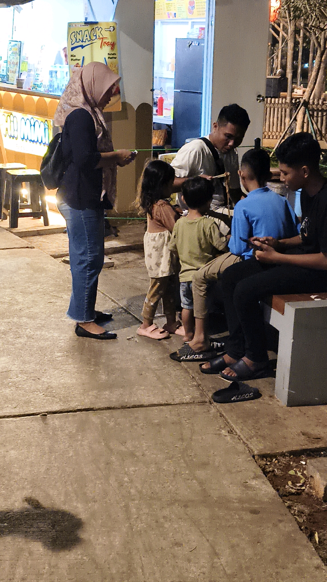 The image shows a group of people standing and sitting in an outdoor setting, possibly a food stall or a public area, in West Jakarta, Indonesia. In the foreground, a woman wearing a pink hijab and blue jeans stands near a food stall with a sign that reads "SNACK Tray." She appears to be interacting with a man standing next to her, who is wearing a white t-shirt and is holding a stick with food.
Several children are also present. Two young girls in casual clothing are standing near the woman and the man, and a boy wearing a green shirt and brown pants is standing next to them. Another boy in a blue t-shirt is sitting on a bench, looking at his phone. To the right of the bench, a man in a black t-shirt is also sitting.
The ground is made of concrete slabs, and there is some dirt and vegetation visible on the side. The background features parts of the food stall, including a refrigerator and a menu board, as well as some decorative elements that appear to be part of the establishment. The lighting suggests it is evening or nighttime. The people are dressed in casual attire, consistent with a relaxed, public gathering.
