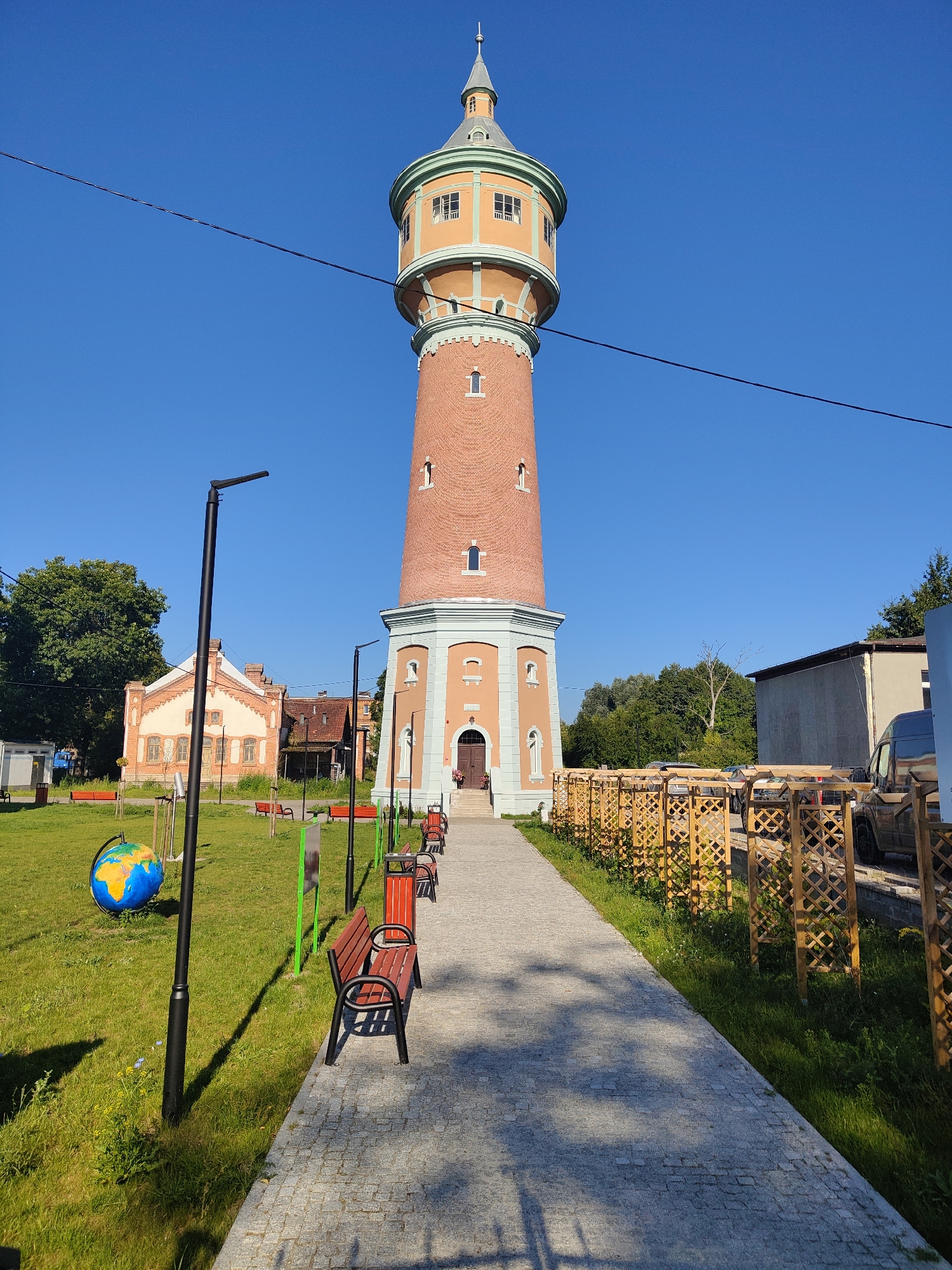 The image shows a tall, brick water tower in Drezdenko, Poland. The tower has an orange section near the top with windows and a green roof. A path made of cobblestones leads to the entrance of the tower, where there are two wooden doors. Benches are placed along the path. To the left of the path is a grassy area with a large globe decoration, streetlights, and more benches. In the background, there are several buildings, including a light-colored building with a red roof. Trees are visible behind the buildings and along the right side of the image. A wooden fence runs along the right edge of the path, separating it from a grassy area where cars are parked. The sky is clear and blue. There are no people visible in the image.