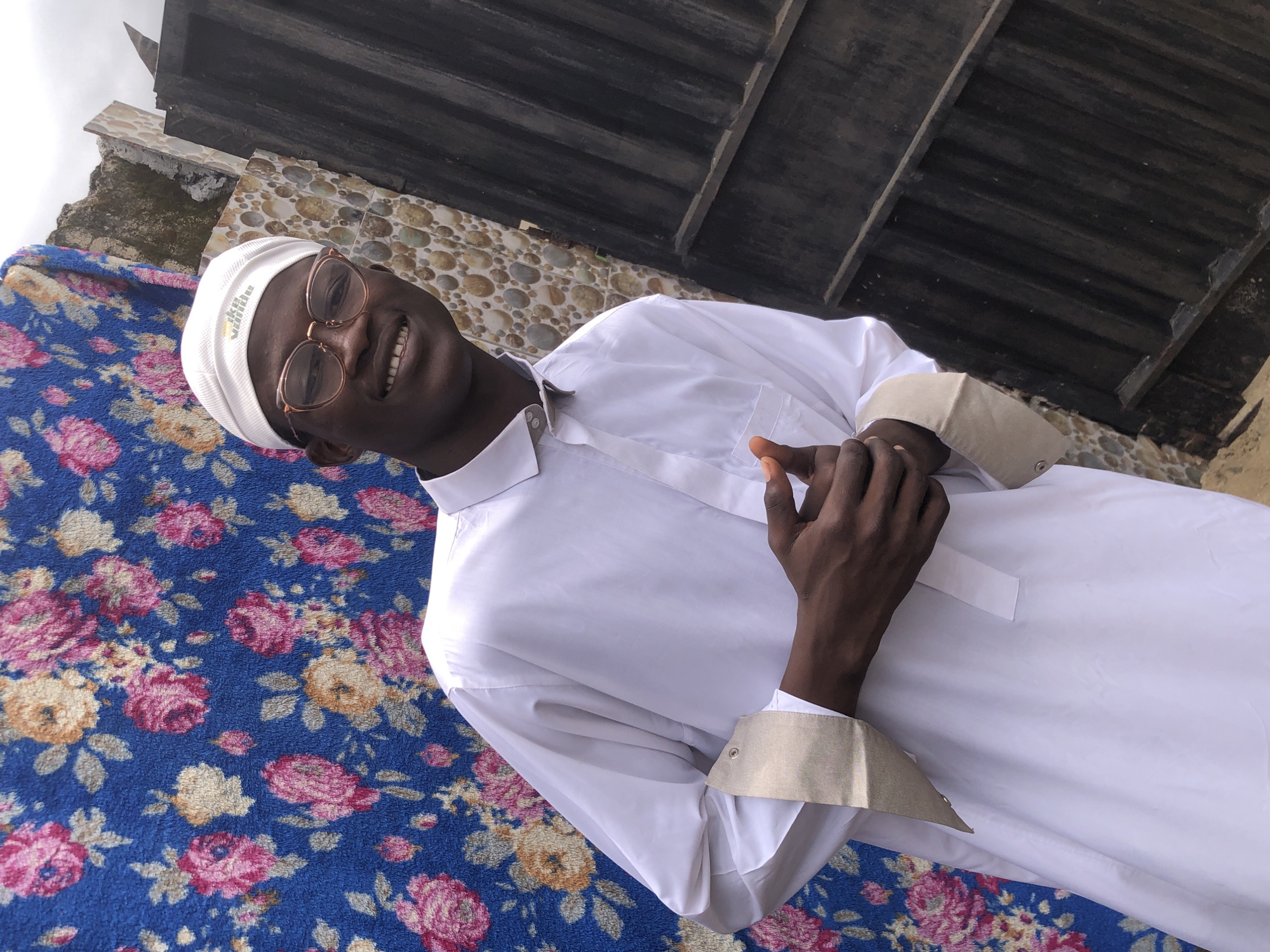 A medium shot captures a young Black male wearing a white kufi and white traditional attire, with the sleeves cuffed with gold fabric, standing outdoors. He is smiling and looking towards the camera, with his hands clasped in front of him. The background features a blue floral tapestry on the left and a dark metal gate on the right. A textured wall with round, beige and brown tiles is visible between the tapestry and the gate. The scene is outdoors, likely in a residential area of Ado, Nigeria. The lighting suggests it is daytime, with diffused light creating soft shadows. The image is taken from a slightly low angle.