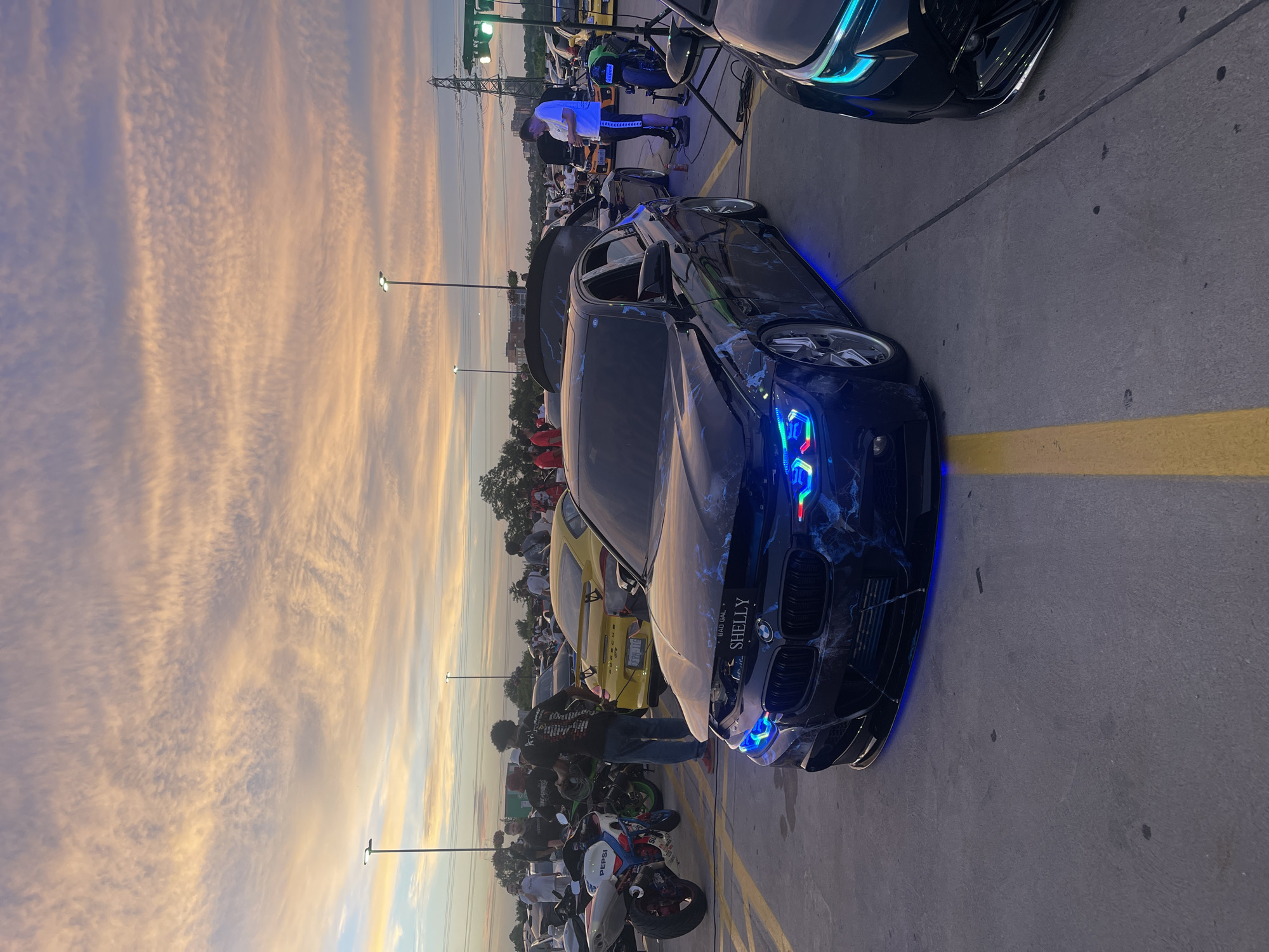 This is a medium shot, eye-level, horizontal, outdoor photograph taken at dusk in a parking lot, likely a car meet. The sky is filled with wispy, striated clouds in shades of pink, orange, and light blue, indicative of sunset. Two streetlights and a distant power transmission tower are visible against the sky.
In the foreground, a black BMW sedan with custom blue and white marble-like paintwork and underglow lighting is the primary subject. The hood is open, revealing the engine bay, and the car has a custom front bumper. A license plate on the front of the car reads "BAD GAL SHELLY". The car is parked on asphalt marked with yellow lines.
To the left of the BMW, a white and red motorcycle with "PEPSI" branding is parked. Behind the motorcycle, a yellow Porsche 911 is visible. Further back, a crowd of people are gathered around various vehicles, including other cars and motorcycles. Some individuals are standing and observing, while others appear to be interacting with the vehicles. The overall atmosphere suggests a casual car enthusiast gathering. The presence of streetlights and the architecture of the distant buildings suggest an urban or suburban setting. The location is identified as being in the city of Toronto, Canada.