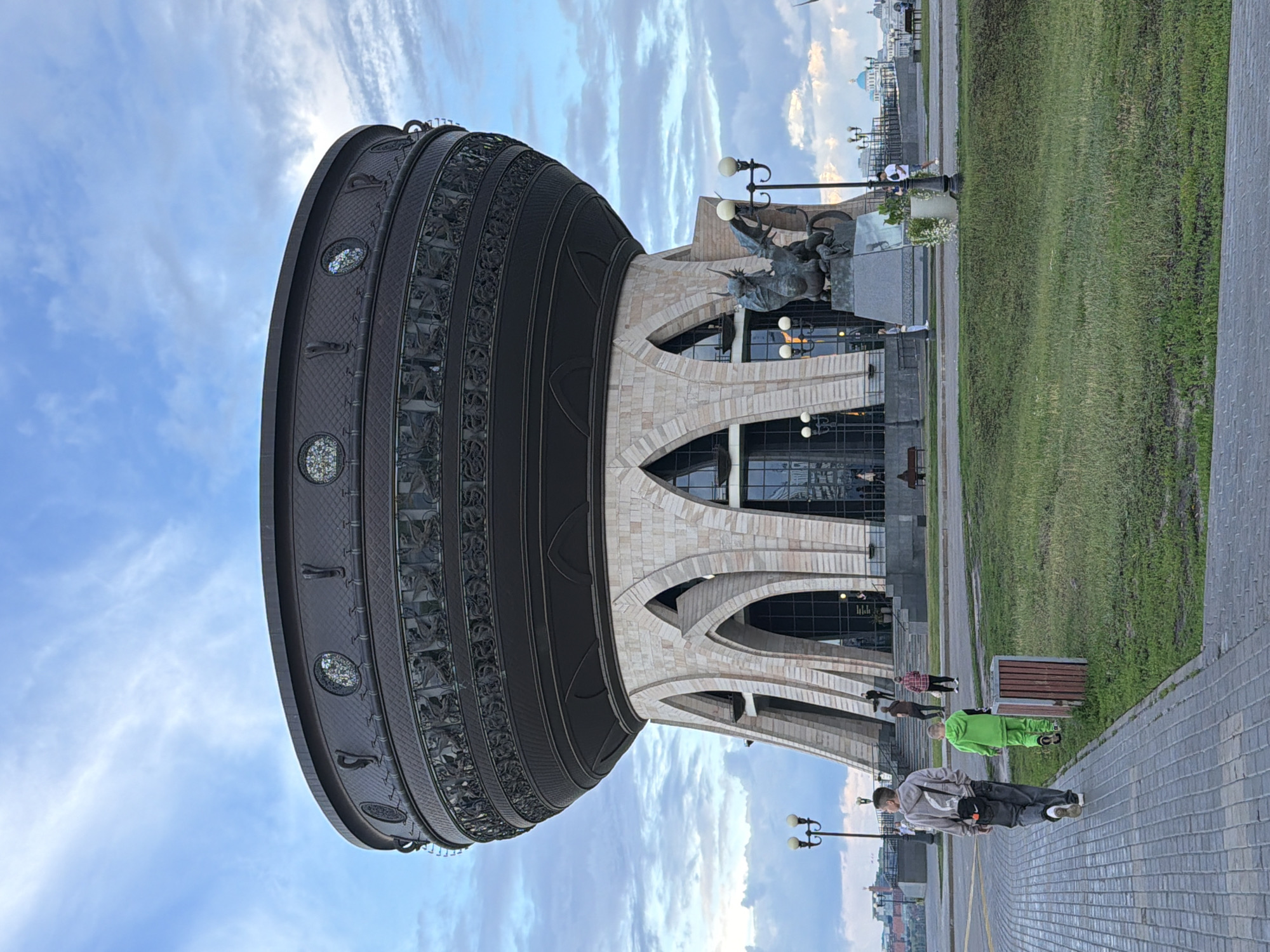 The image shows a street view in Kazan, Russia. The main subject is a large, cylindrical, dark-colored monument with intricate metalwork and decorative circular elements. To the right of the monument is a modern building with a facade of light-colored stone and large arched windows. In front of the building, a sculpture of a figure on horseback is visible.
The scene is an outdoor public space with a paved walkway and a well-maintained grassy area. Several people are present in the image. In the foreground, a person in a grey hoodie and black pants is walking on the paved path. Further back, a person in a bright green tracksuit is standing on the path. Other individuals are scattered throughout the scene, some walking and others appearing to be standing near the building.
The sky is partly cloudy with a mix of blue sky and white clouds, suggesting it might be daytime with pleasant weather. The lighting indicates it could be late afternoon or early evening, with the sun casting soft light. The overall atmosphere appears to be calm and ordinary, with people going about their day in a public urban setting. There is no visible text in the image.