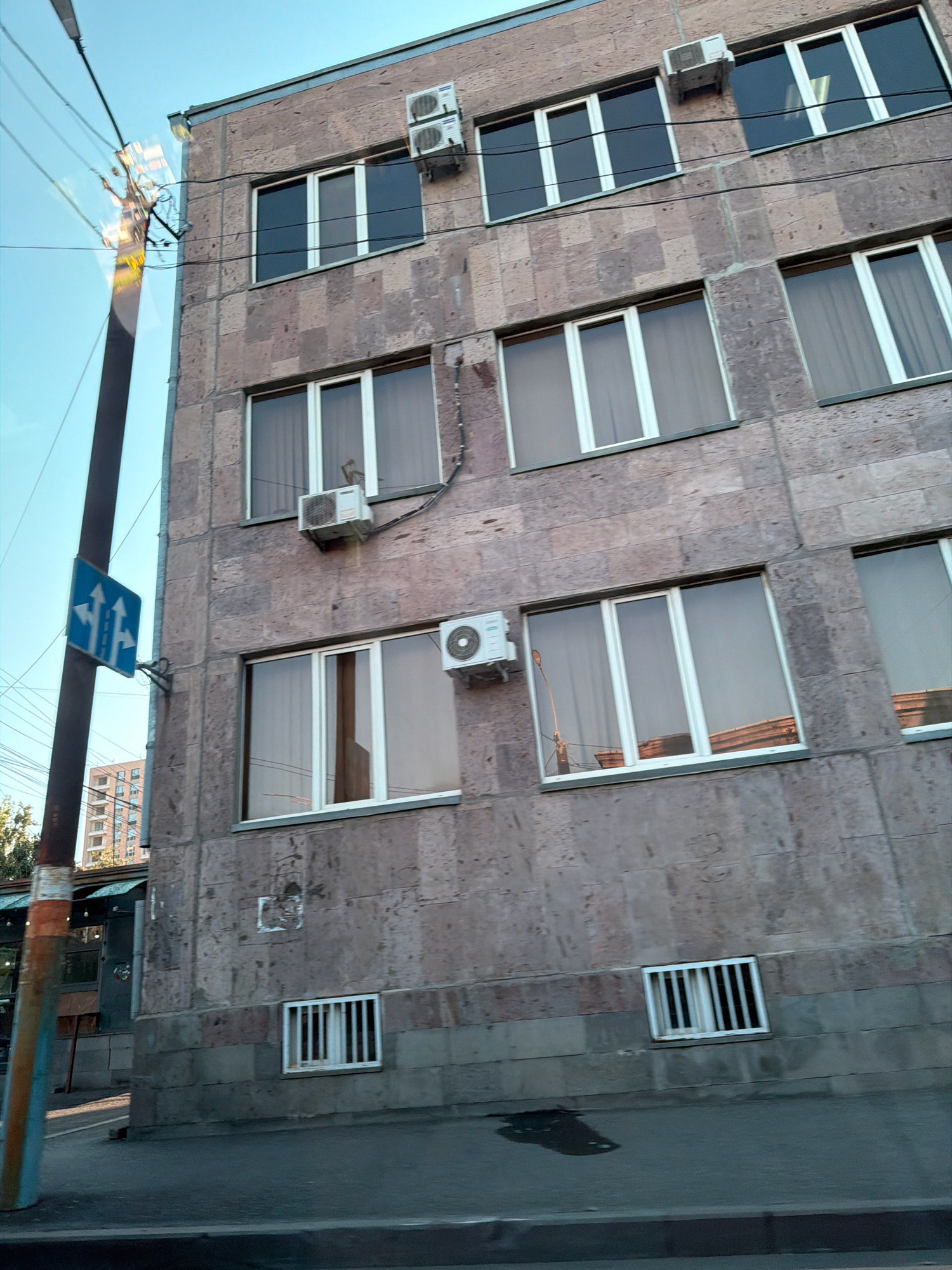 The image shows a side view of a multi-story building, likely an apartment building or office complex, with a stone facade and several windows. The building appears to be located on a street in Yerevan, Armenia, as indicated by the architecture and the presence of a traffic sign with Armenian script. The sky is clear and blue, suggesting it is daytime.
The building has a somewhat dated architectural style. The facade is made of stone blocks in varying shades of brown and gray. Multiple windows are visible on the side of the building, most of them rectangular and framed in white. Some windows have sheer curtains visible inside, while others are darker, reflecting the sky or interior lights.
Several air conditioning units are mounted on the exterior walls of the building, indicating a possibly warm climate.
In the foreground, a street or sidewalk is visible, with a dark asphalt surface. A portion of a utility pole is on the left side of the frame, with electrical wires extending from it. Attached to this pole is a blue traffic sign with white arrows, indicating traffic flow or lane designations.
In the background, another building with a lighter facade and multiple windows can be seen peeking out from behind some trees.
The overall scene is urban and functional, typical of a residential or commercial area in a city like Yerevan. There are no people or animals visible in the image. The image appears to have been taken from a moving vehicle, given the slight tilt of the building and the dynamic perspective.