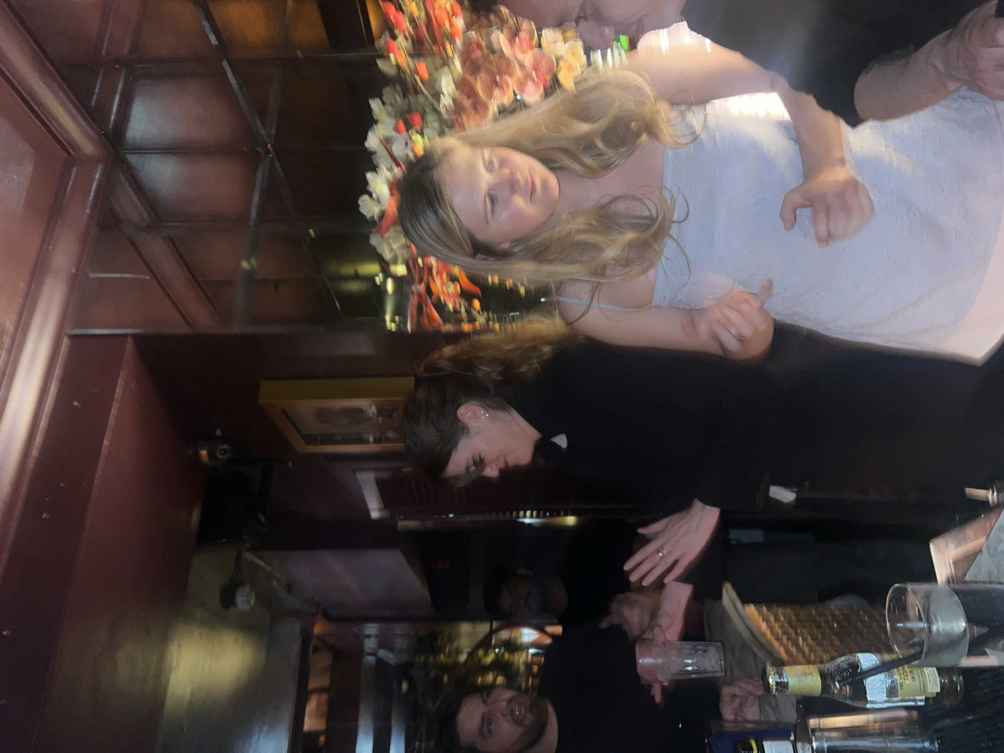 A scene inside what appears to be a restaurant or bar in London, United Kingdom. A woman in a white dress, possibly a bride, stands near a bar counter. She has long blonde hair and is wearing a floral headpiece. Two people are interacting with the woman: one seems to be holding her hand (upper right corner) while the other is a bartender in black, who is standing behind the bar. The bar counter has various bottles, glasses, and bar equipment on it. The setting has warm, dimly lit interiors with ornate decor, including what looks like mirrored ceilings or walls with geometrical patterns. The overall atmosphere seems lively.
