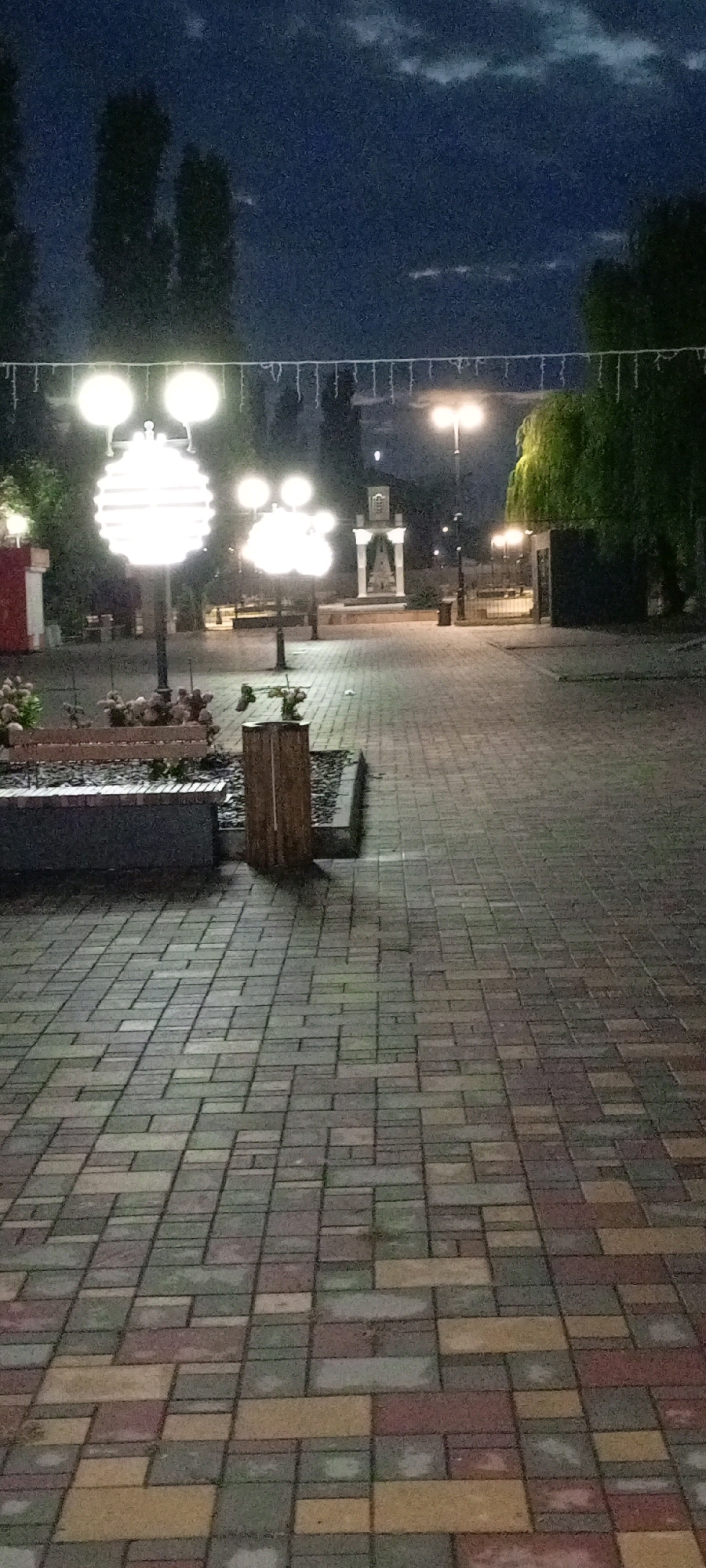 The image depicts a nighttime scene in a city park or public square in Kirsanov, Russia. The foreground is dominated by a paved walkway made of multi-colored rectangular bricks. To the left, a low stone bench is visible, bordered by dark mulch and some sparse, low-lying plants. Next to the bench, a wooden trash receptacle stands.
Further into the scene, a pathway continues, lined with lampposts that emit a bright, warm light. Two prominent lampposts feature multiple spherical lights, one of which has a large, orb-like decorative element. In the background, a small, white architectural structure, possibly a monument or gazebo, stands illuminated. Tall, dark trees are visible on either side of the background, with dense foliage on the right. The sky above is dark blue with scattered clouds, suggesting dusk or nighttime. Decorative string lights are visible spanning across the top of the frame, adding to the festive atmosphere. No people are present in the image.