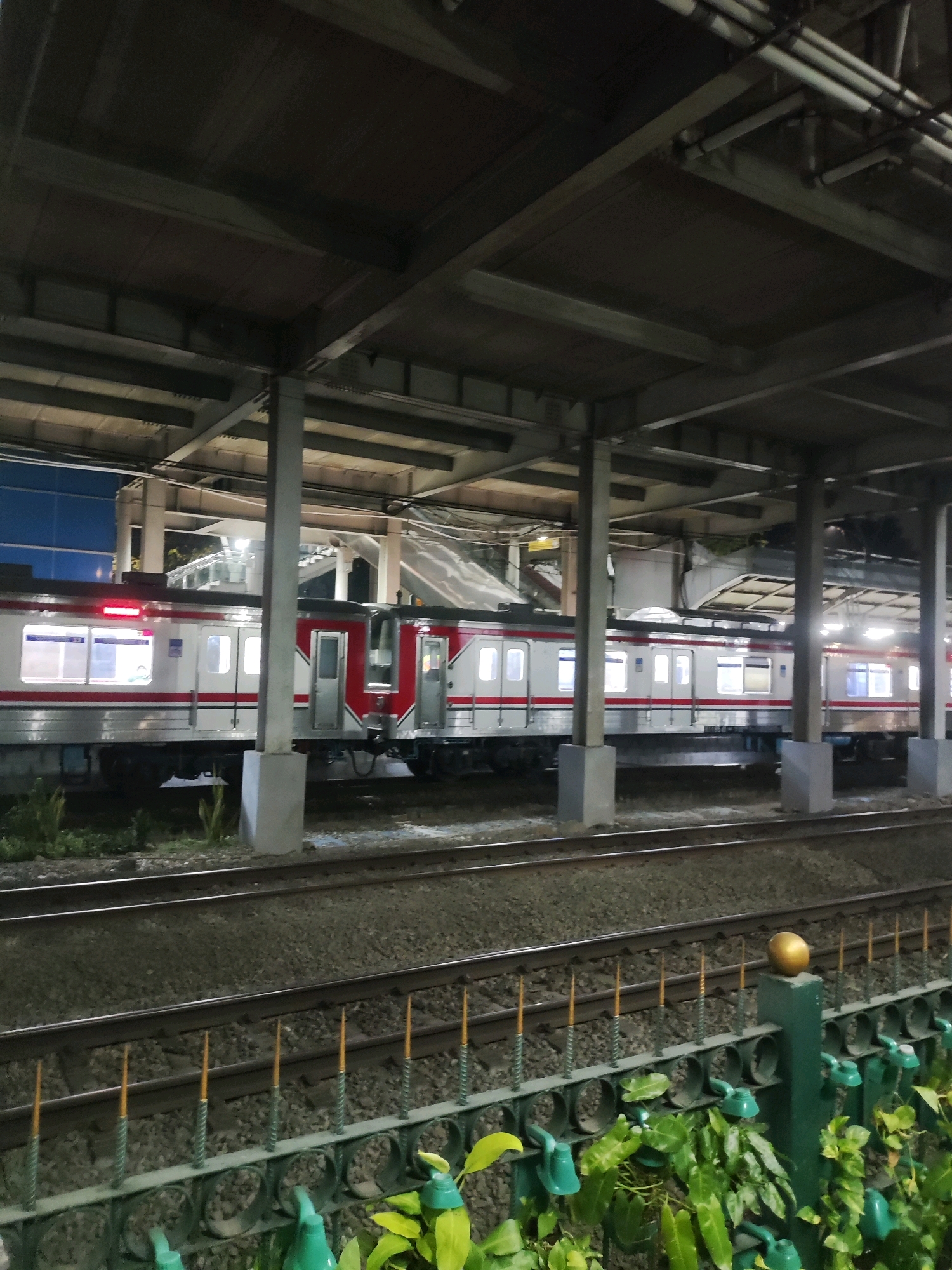 The image captures a nighttime scene at a train station in East Jakarta, Indonesia. A white and red train with illuminated windows is stopped on the tracks. The station features a covered platform supported by concrete pillars. In the foreground, a green fence with pointed metal spikes and decorative circular openings lines the area, with lush green plants growing around it. The tracks are visible in the mid-ground, and beyond them, part of a blue building can be seen. The overall lighting suggests it is after sunset, with artificial lights illuminating the platform and train.