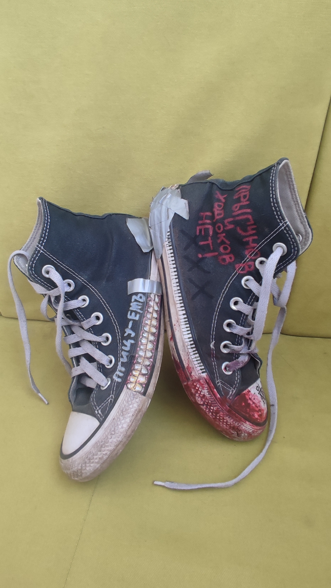 Two black high-top sneakers are positioned on a yellow-green surface. The right sneaker has red markings on the toe and sole, resembling blood splatter, and red Cyrillic writing on its side that states "Присутствие ходоков НЕТ!", which translates to "The presence of walkers is NO!". The left sneaker displays a design that mimics a set of teeth on the sole's edge, accompanied by illegible Cyrillic text. Silver tape is applied to both sneakers at different points. The laces of both shoes are grey. The background surface appears to be a textured fabric, like a couch. The photograph was taken in Shelekhov, Russia.