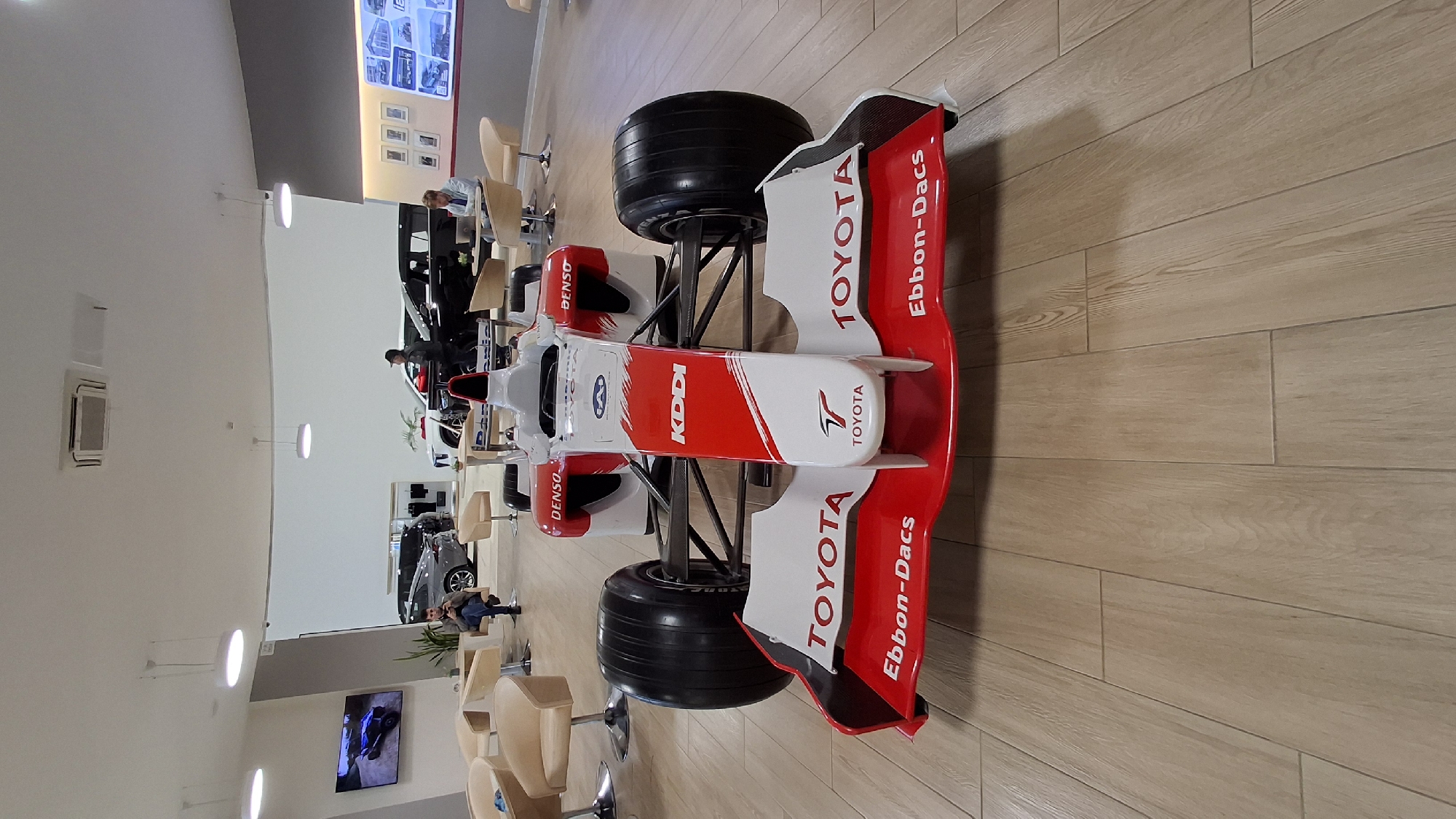 This is a photograph of a Toyota Formula 1 race car model displayed indoors, likely in a showroom or exhibition space. The car is predominantly white with red accents and features sponsor logos such as "TOYOTA," "DENSO," "KDDI," and "Ebbon-Dacs." The setting has light-colored wooden plank flooring.
In the background, several people are visible. Some are standing near other vehicles, possibly cars, while others are seated in chairs. One person is leaning over a black car. There's a television screen mounted on the wall displaying an image. The overall lighting suggests it is daytime, and there are no obvious cues about the weather. The atmosphere appears casual, with people browsing or waiting. The location is identified as Novogorelovo, Russia.