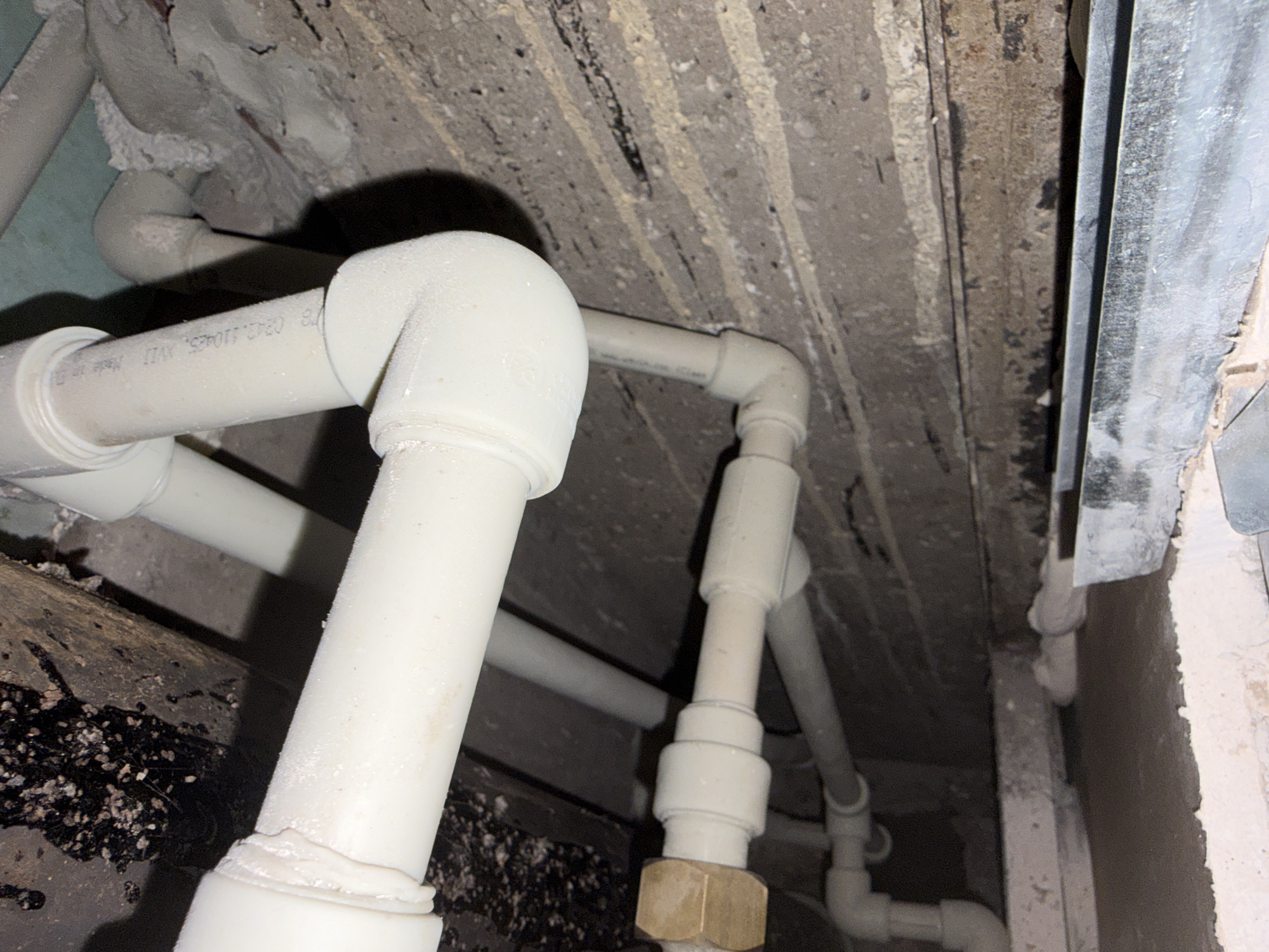 The image depicts a close-up, low-angle view of plumbing pipes running along a rough, unfinished concrete ceiling. The pipes, primarily made of white plastic, are arranged in various configurations, including elbows and couplings. Some pipes appear dusty or coated with a white residue. The ceiling shows exposed rebar and has a textured, uneven surface. On the left side of the image, a wooden beam with dark, granular debris is visible. In the background, on the right, a metallic duct or pipe runs vertically along a rough, plastered wall. The lighting is dim, casting shadows and highlighting the textures of the materials. There are no people or discernible actions occurring in the scene.