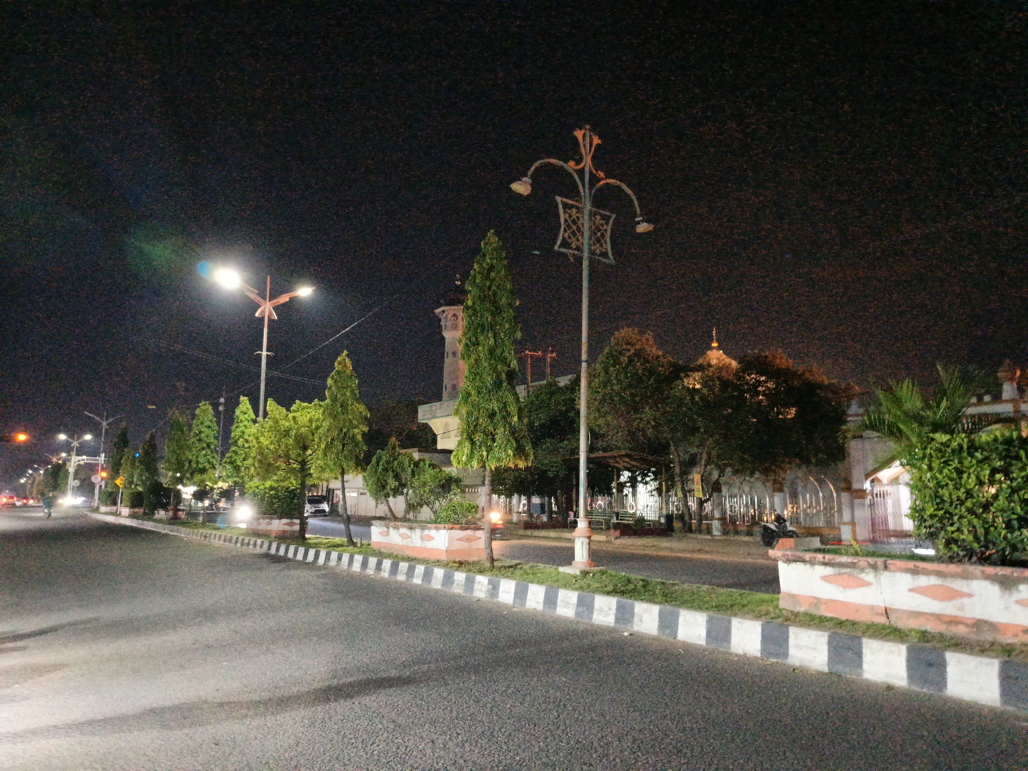 The image captures a night scene on a street in Lhokseumawe, Indonesia. The street is lined with trees and ornamental lamp posts, and a mosque is visible in the background. The road is asphalt, with a distinct divider composed of alternating black and white blocks along the curb. The sky is dark, and the scene is illuminated by streetlights and artificial lighting from the mosque and nearby buildings. There are a few vehicles visible in the distance. The general atmosphere suggests a quiet night in the city.