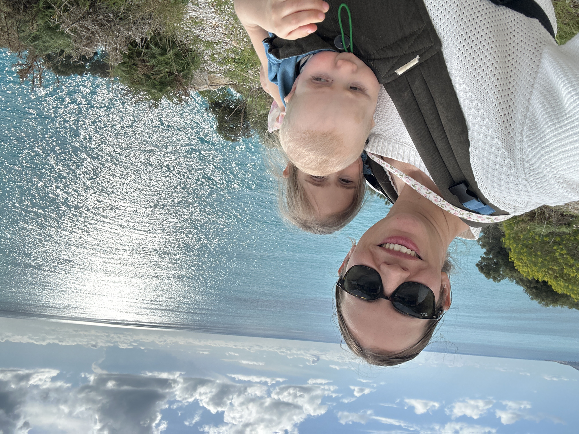 A woman wearing sunglasses and a baby are photographed with a vast body of water and sky in the background. The water shimmers with reflected sunlight. The sky above the water is blue with cumulus clouds. A coastal landscape with vegetation is visible at the top and bottom. The location is likely in Fethiye, Turkey, given the presence of Mediterranean Sea, characteristic topography, and vegetation. The woman appears to be holding the baby in a carrier.